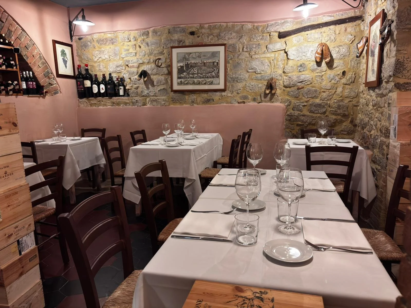 Interior of a cozy Italian restaurant with stone walls, wooden chairs, tables set with white tablecloths, wine glasses, and silverware, decorated with framed pictures, wine bottles, and hanging shoes.