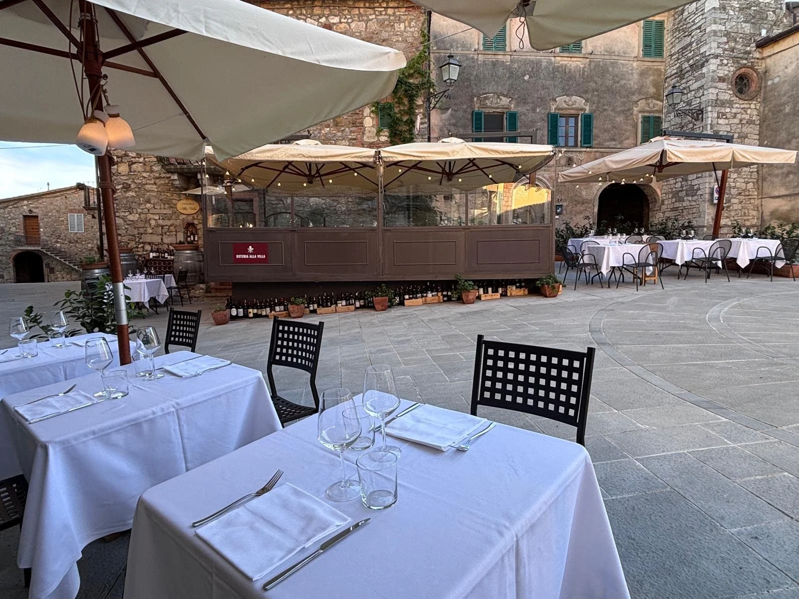 Outdoor restaurant with white tablecloth-covered tables, black chairs, umbrellas, and a view of historic stone buildings and a cobblestone courtyard.