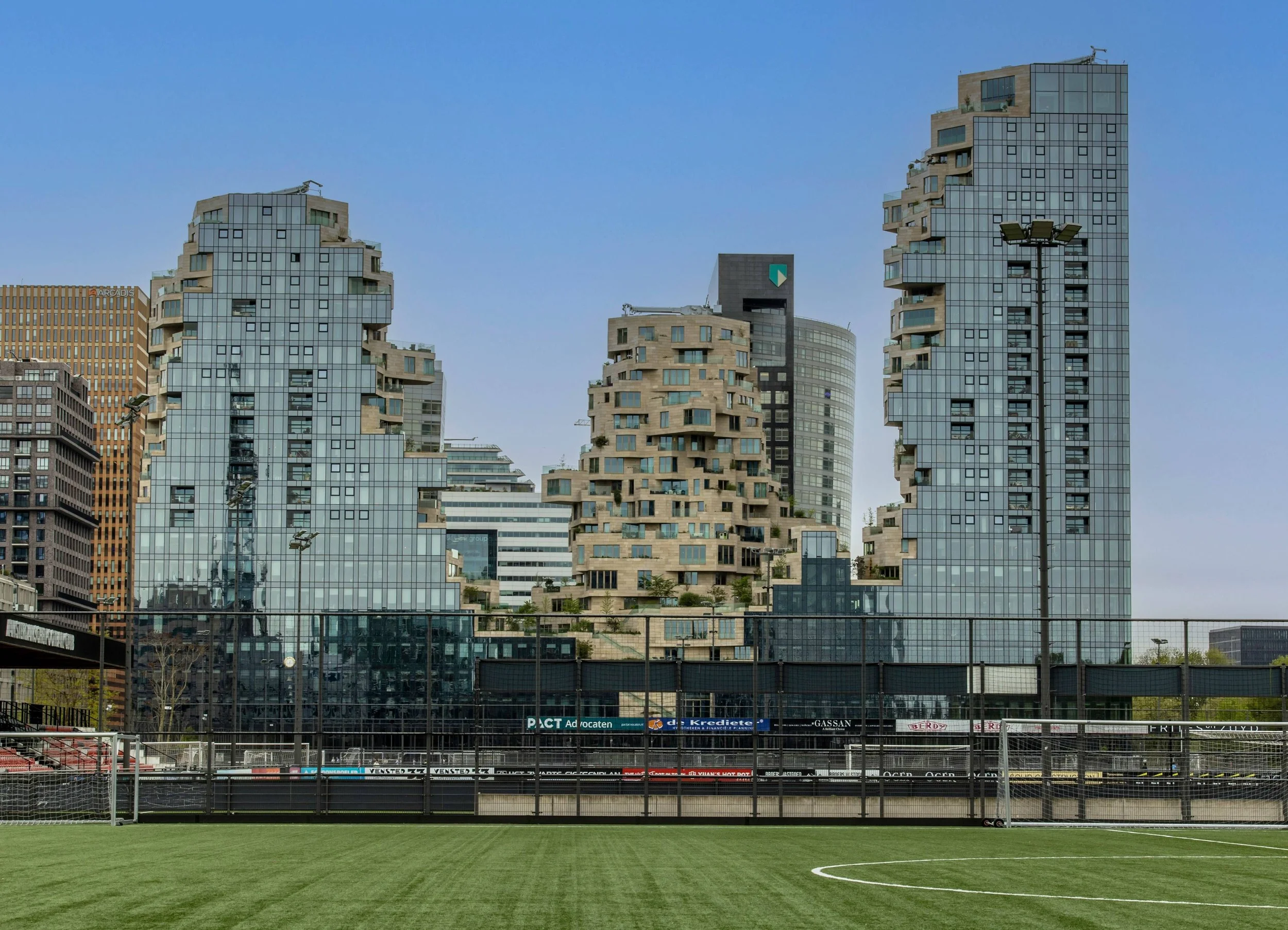 Modern cityscape with high-rise buildings, some with unique stepped designs, viewed from an empty soccer field with a green lawn and a fence.