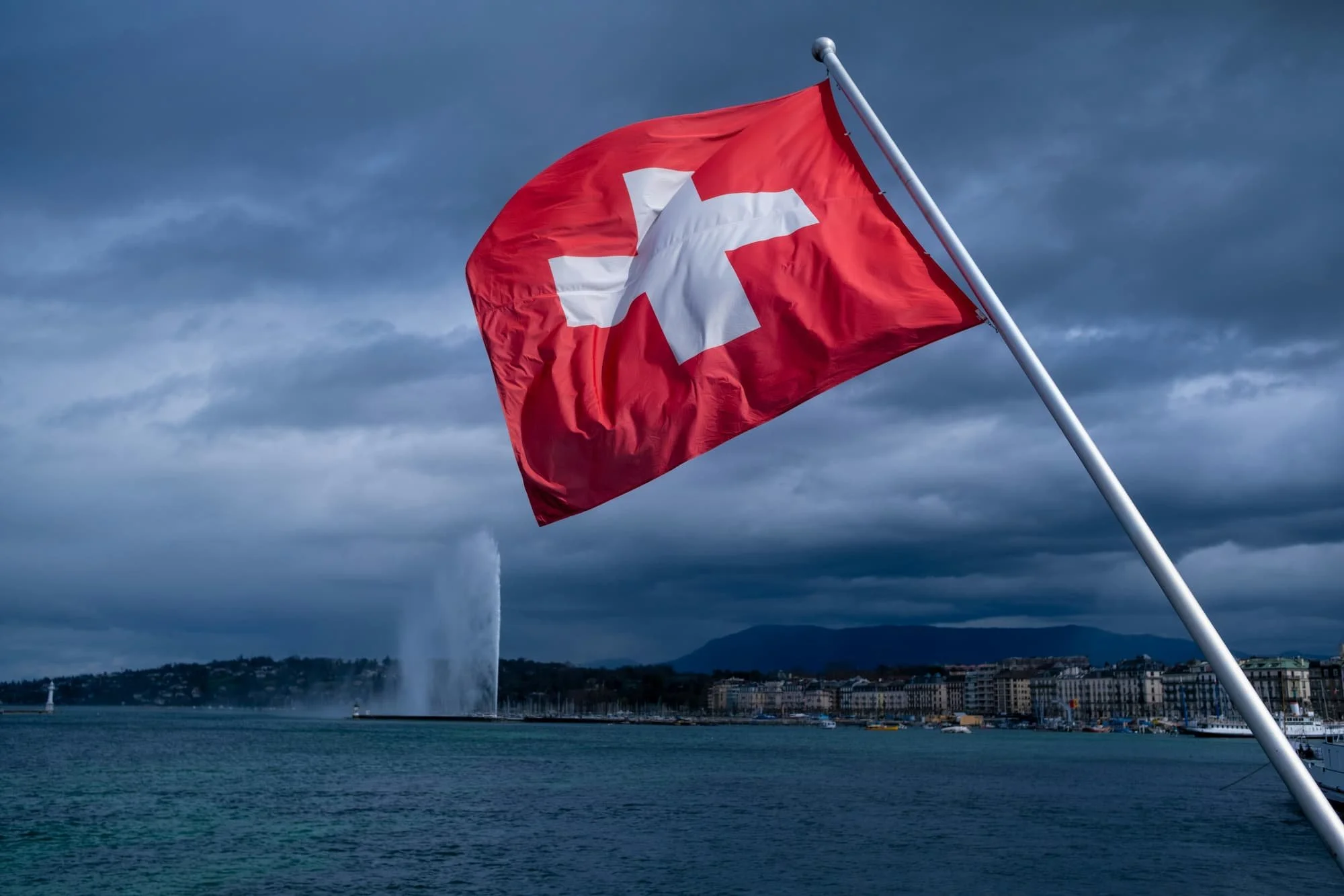 Swiss flag on a flagpole in front of mountainous landscape with cloudy sky.