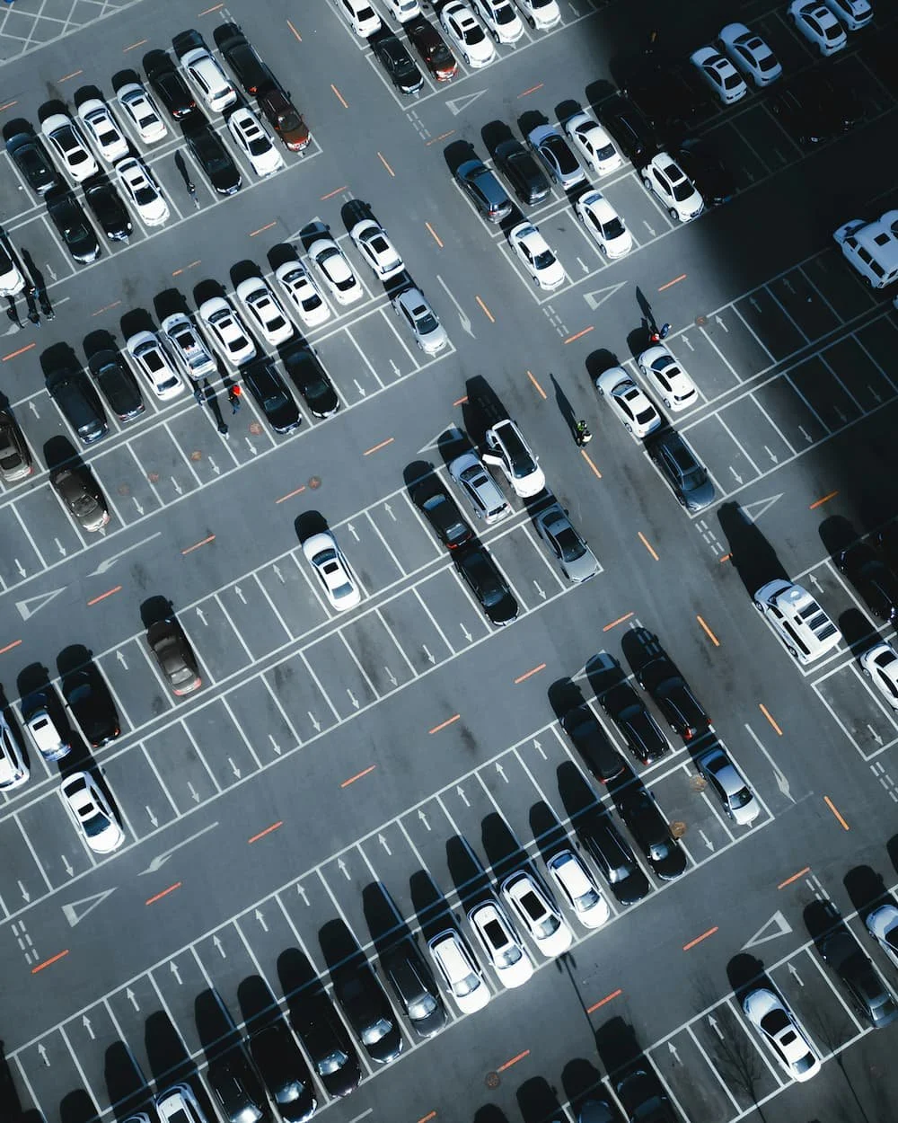 An aerial view of a large parking lot filled with cars, with clearly marked parking spaces and shadows cast by the vehicles.