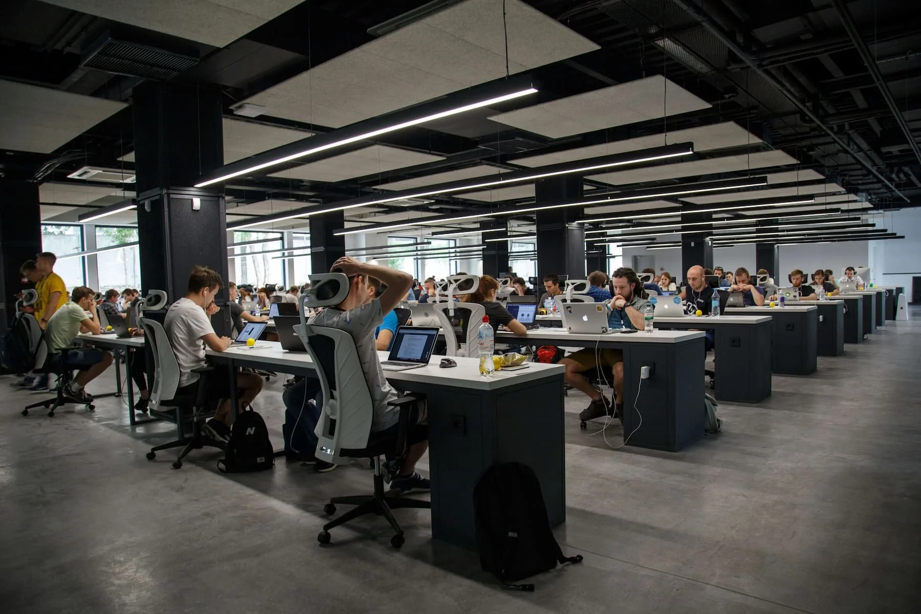 Open office space with several employees working at desks with computers. The room features exposed brick walls, large windows, and ceiling ductwork with wooden beams. There's a mix of office furniture including chairs, sofas, a coffee table, and a large plant in the foreground.