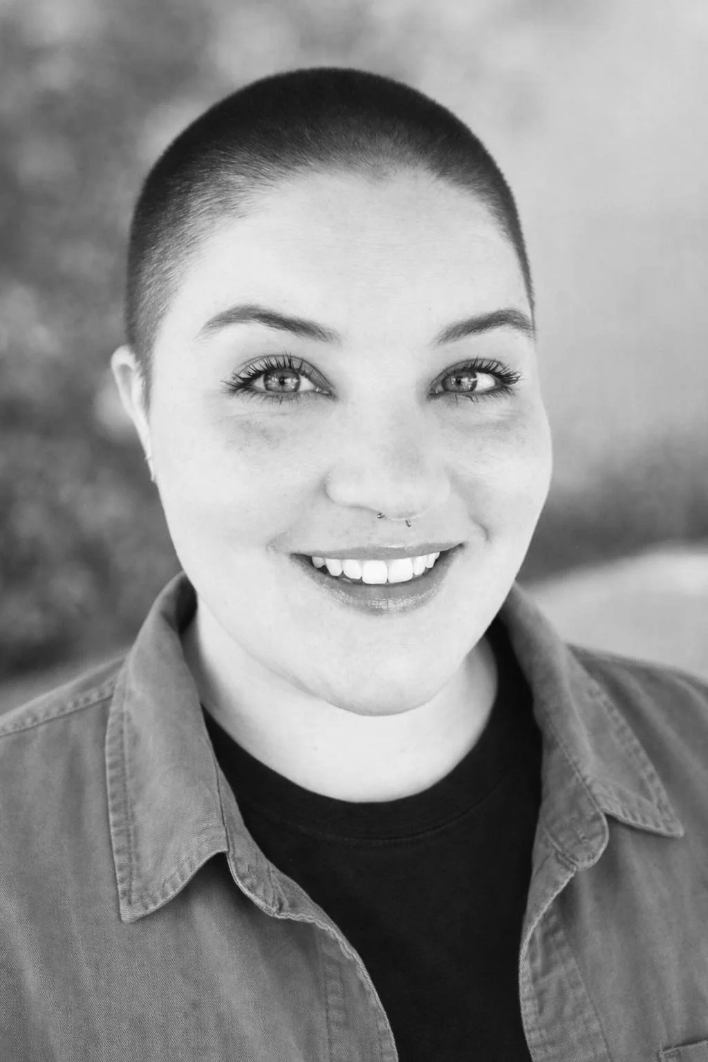 Black and white portrait of a woman with a shaved head, smiling, wearing a jacket and a t-shirt.