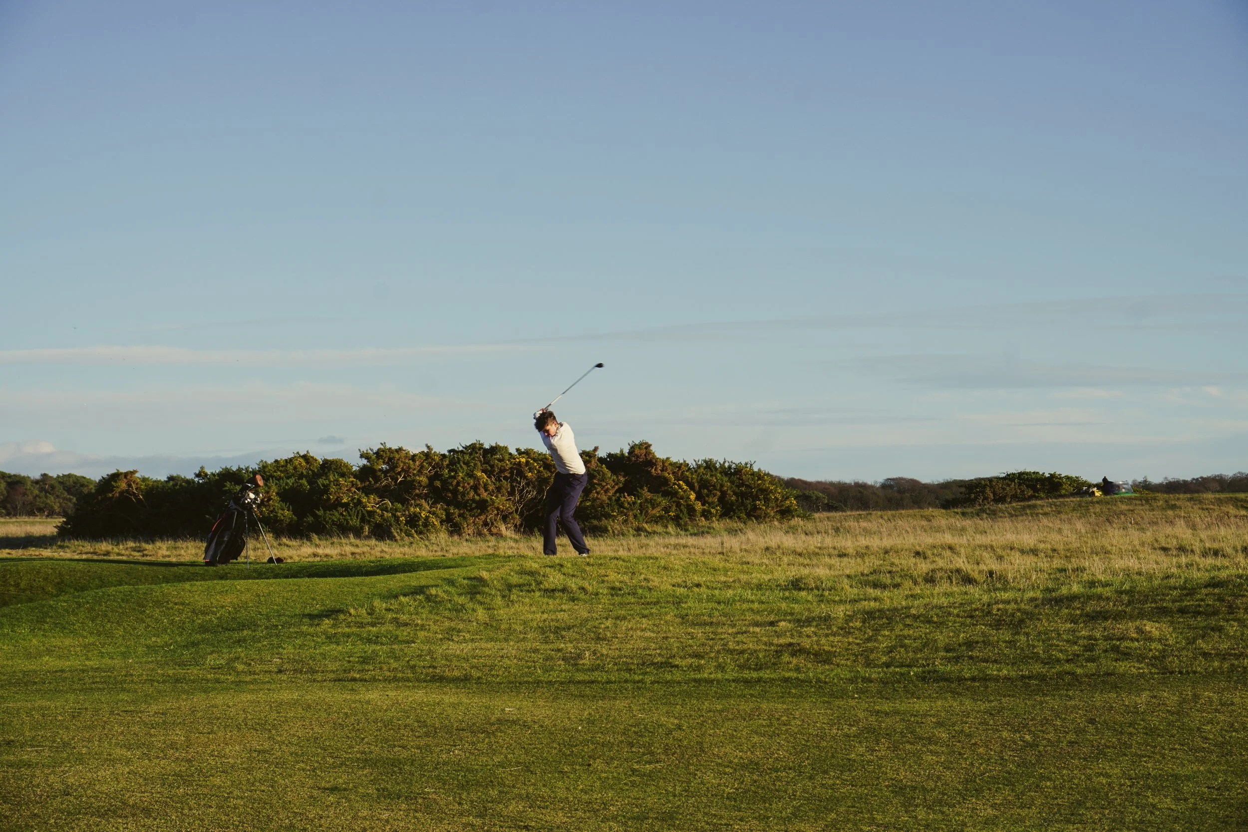 A person playing golf on a sunny day, swinging a golf club on a grassy course with some bushes and a blue sky in the background.