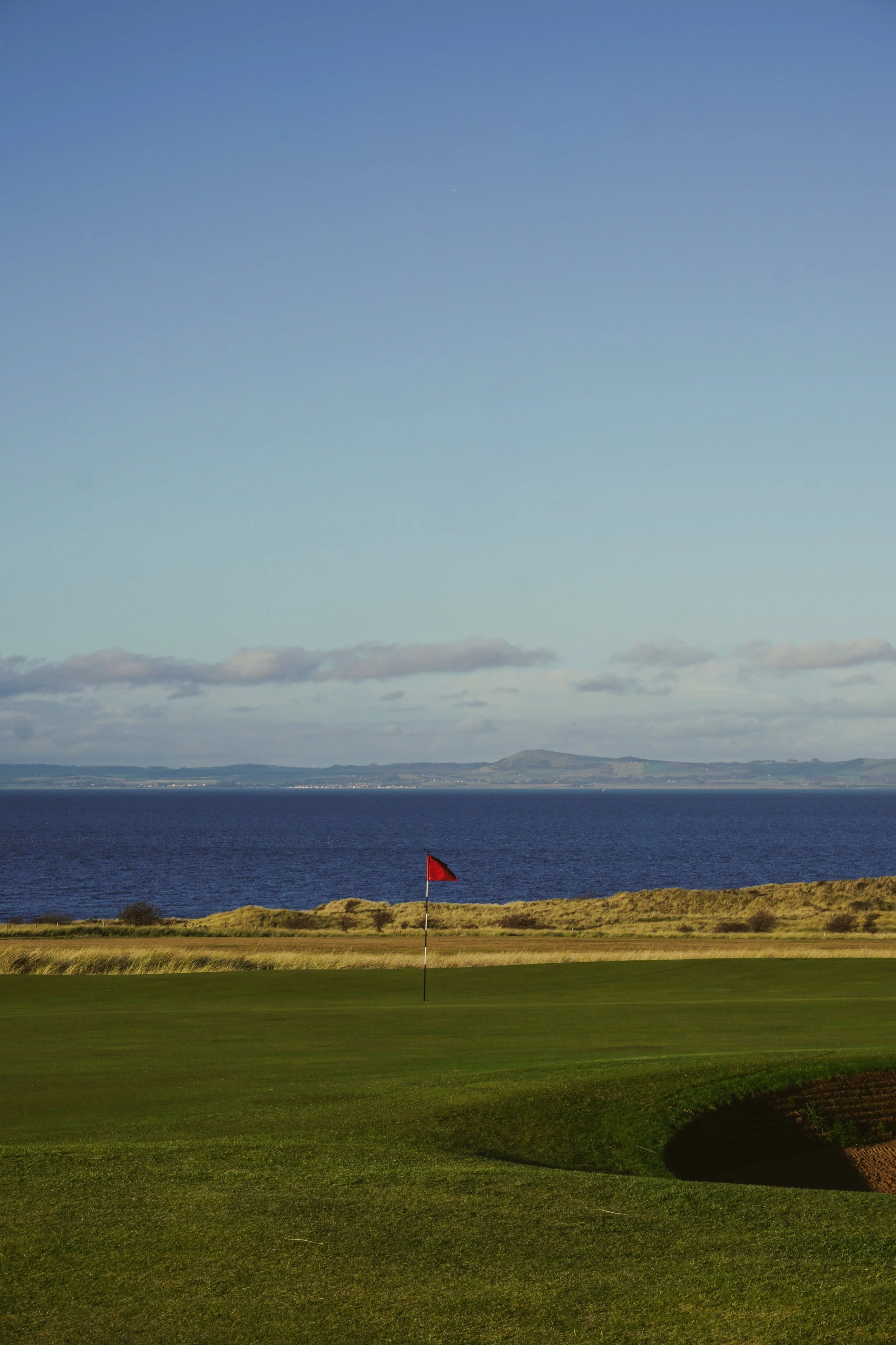A golf course with a green, a red flag, and a view of a large body of water and mountains in the distance.