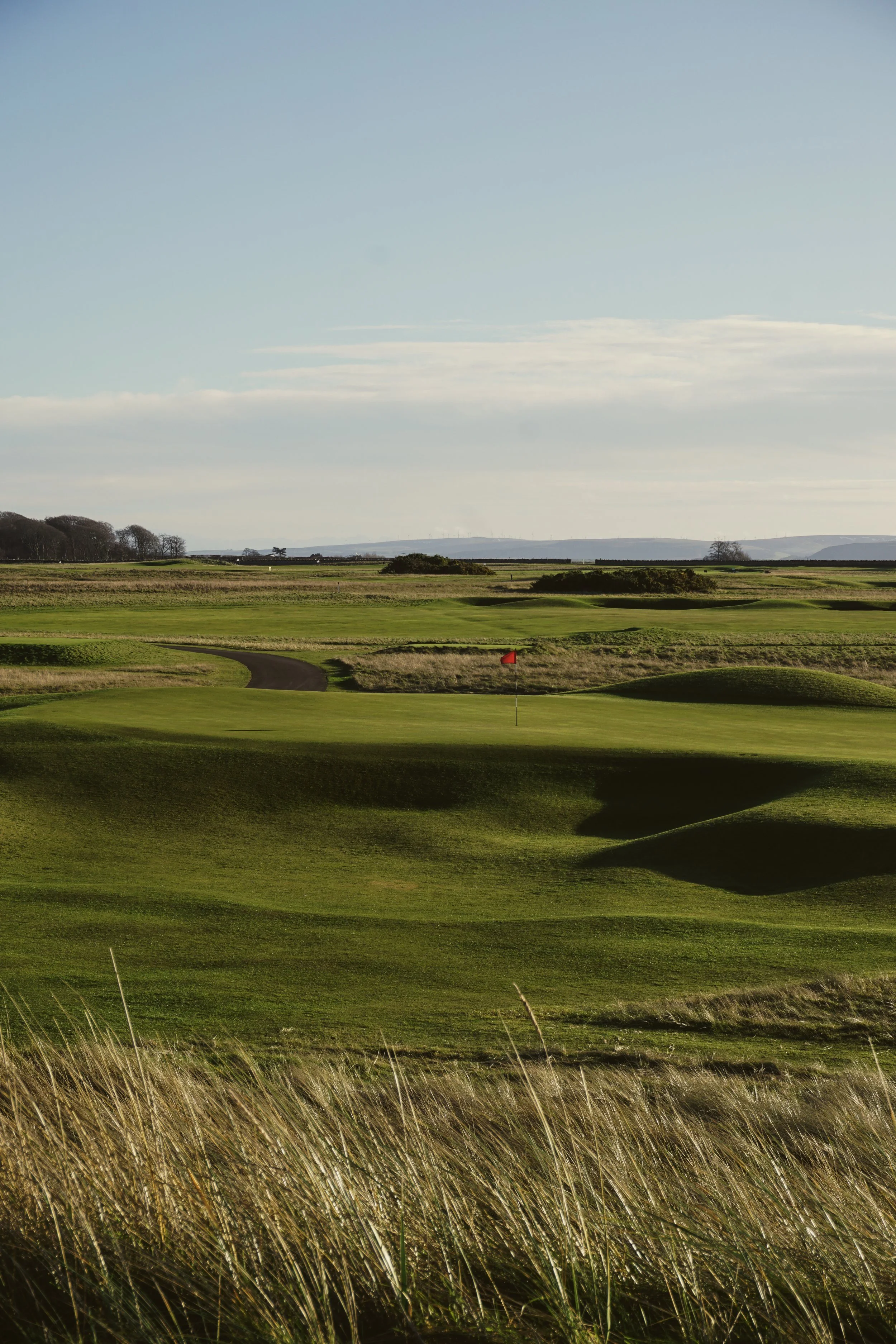 A golf course with green fairways, a flag on the putting green, and rolling terrain under a partly cloudy sky.