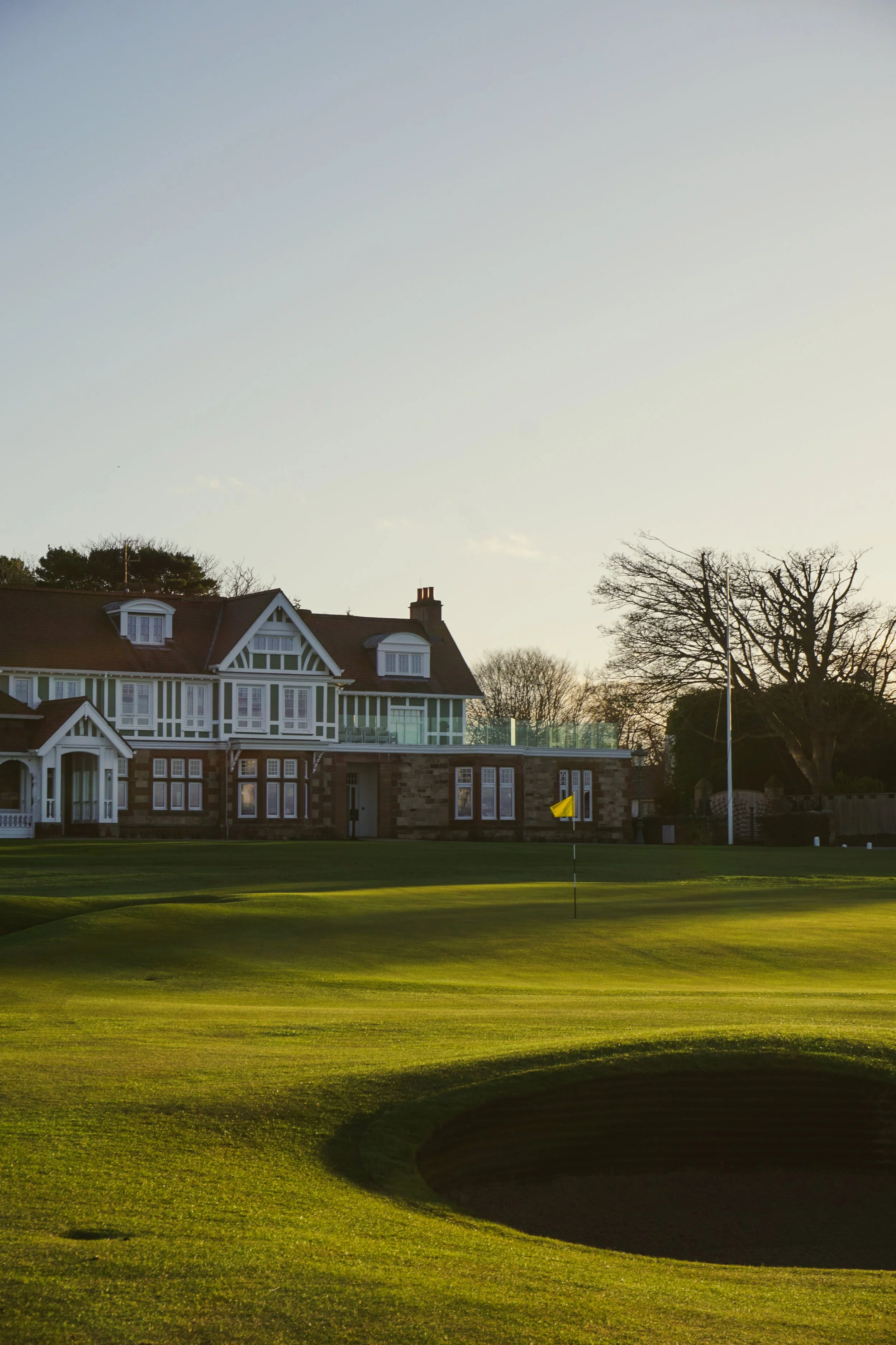 The 18th hole at Muirfield with a flagstick near the middle, adjacent to a large house with multiple windows, a porch, and a gabled roof, during sunset with trees in the background.