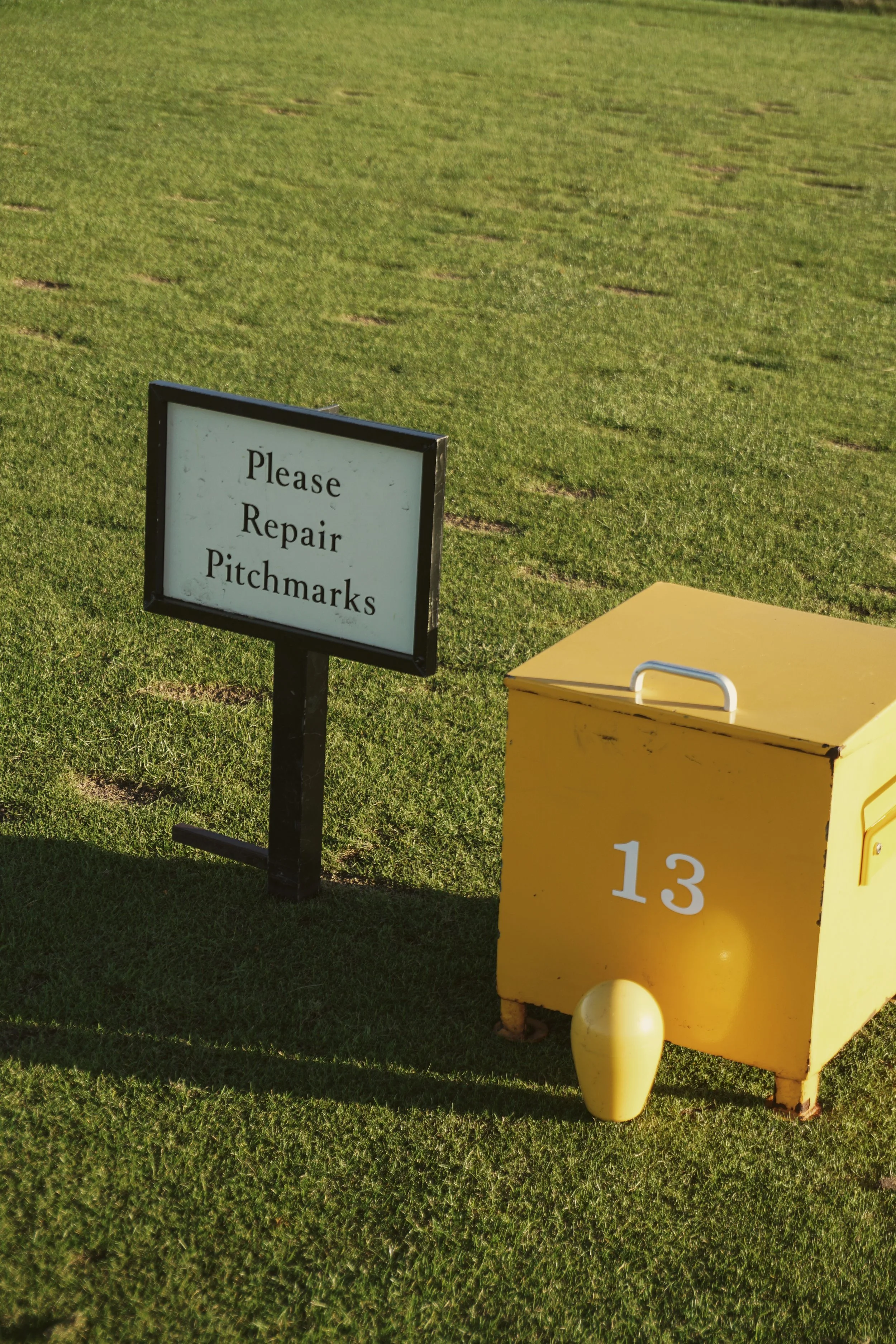 A sign on a golf course that says, "Please Repair Pitchmarks," next to a yellow maintenance cart with the number 13.