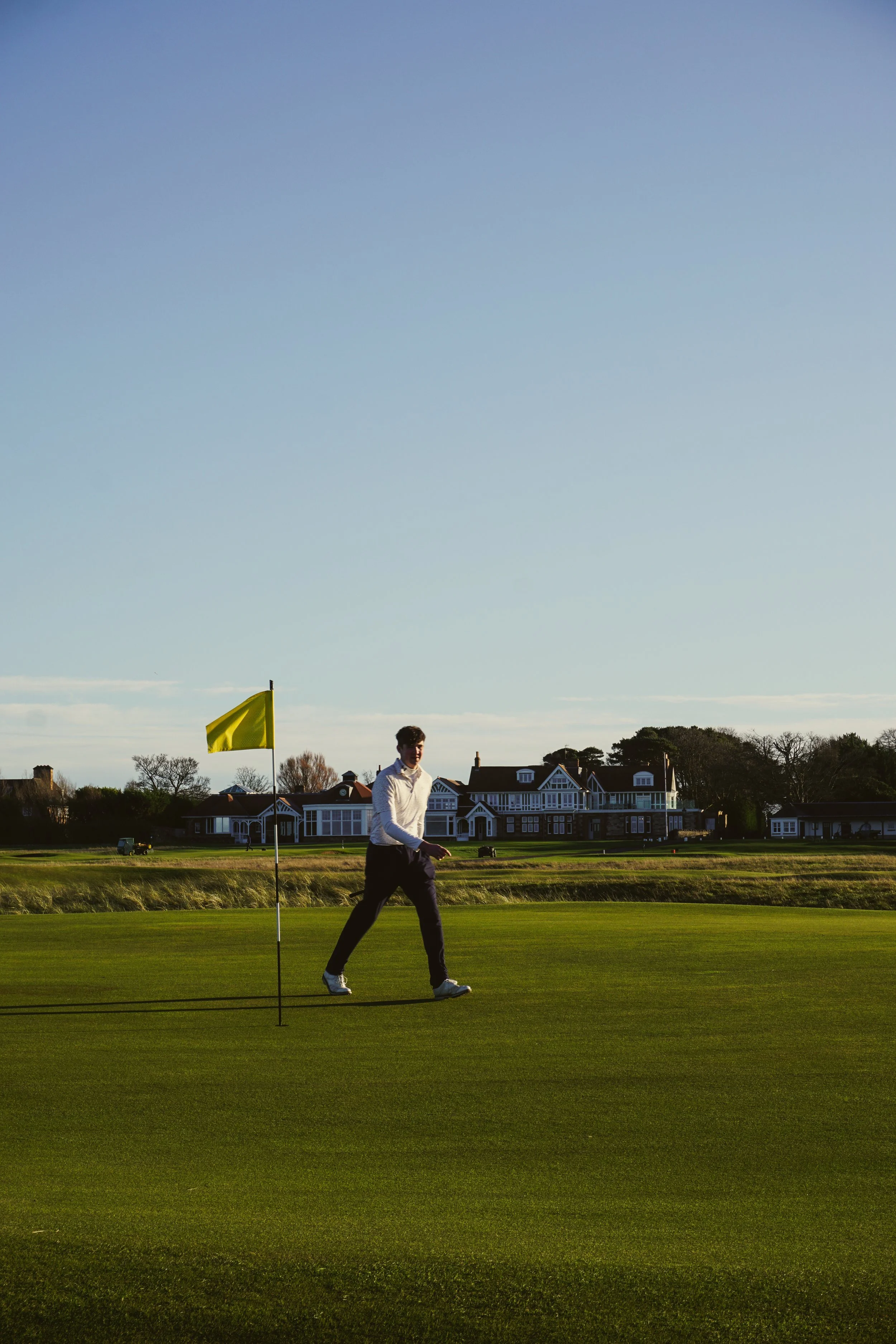 A person walking on a golf course near a yellow flagstick under a clear sky.