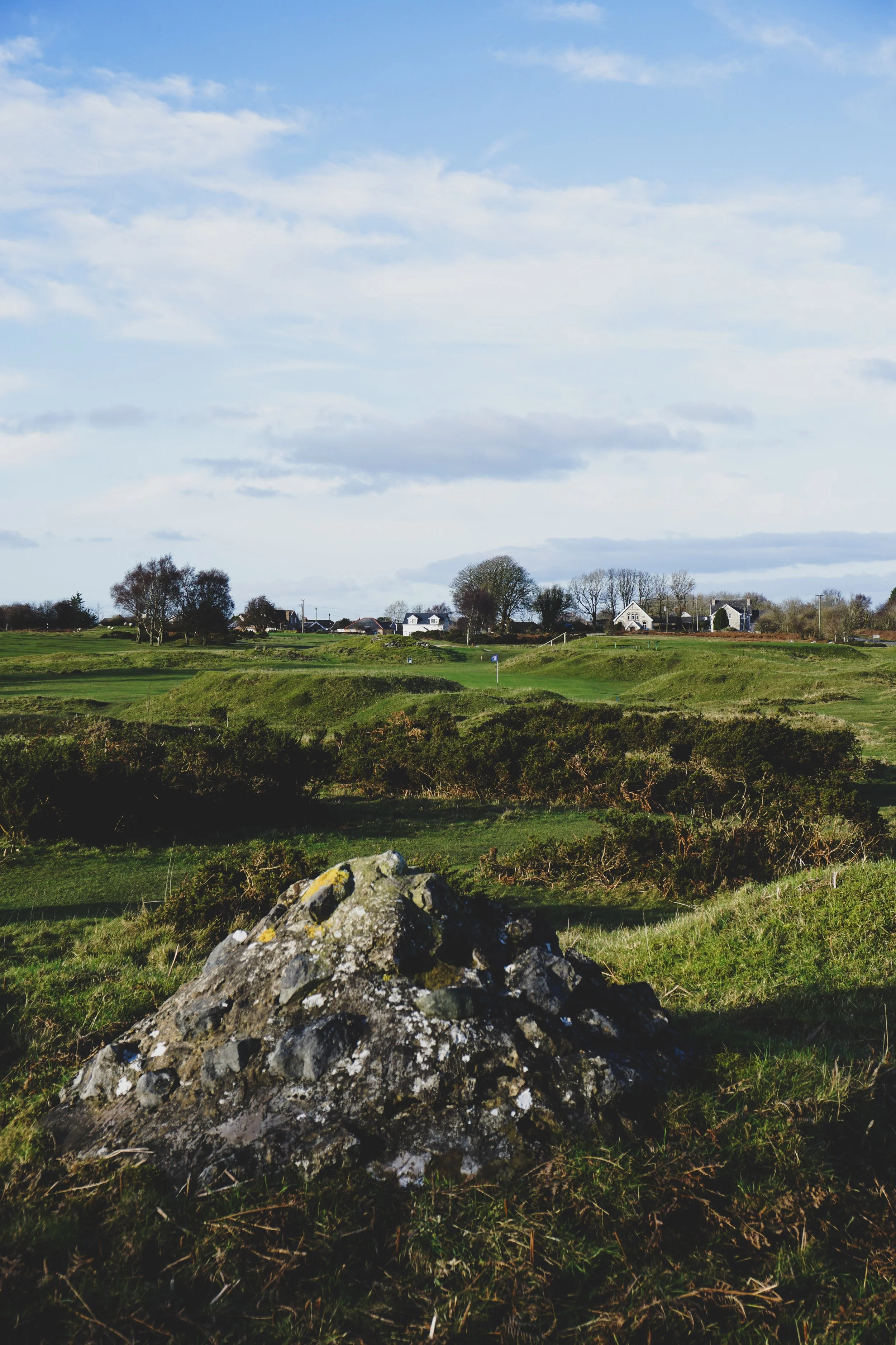 A scenic landscape of a golf course with a large rock in the foreground, green fairways, trees, and houses in the background under a partly cloudy sky.