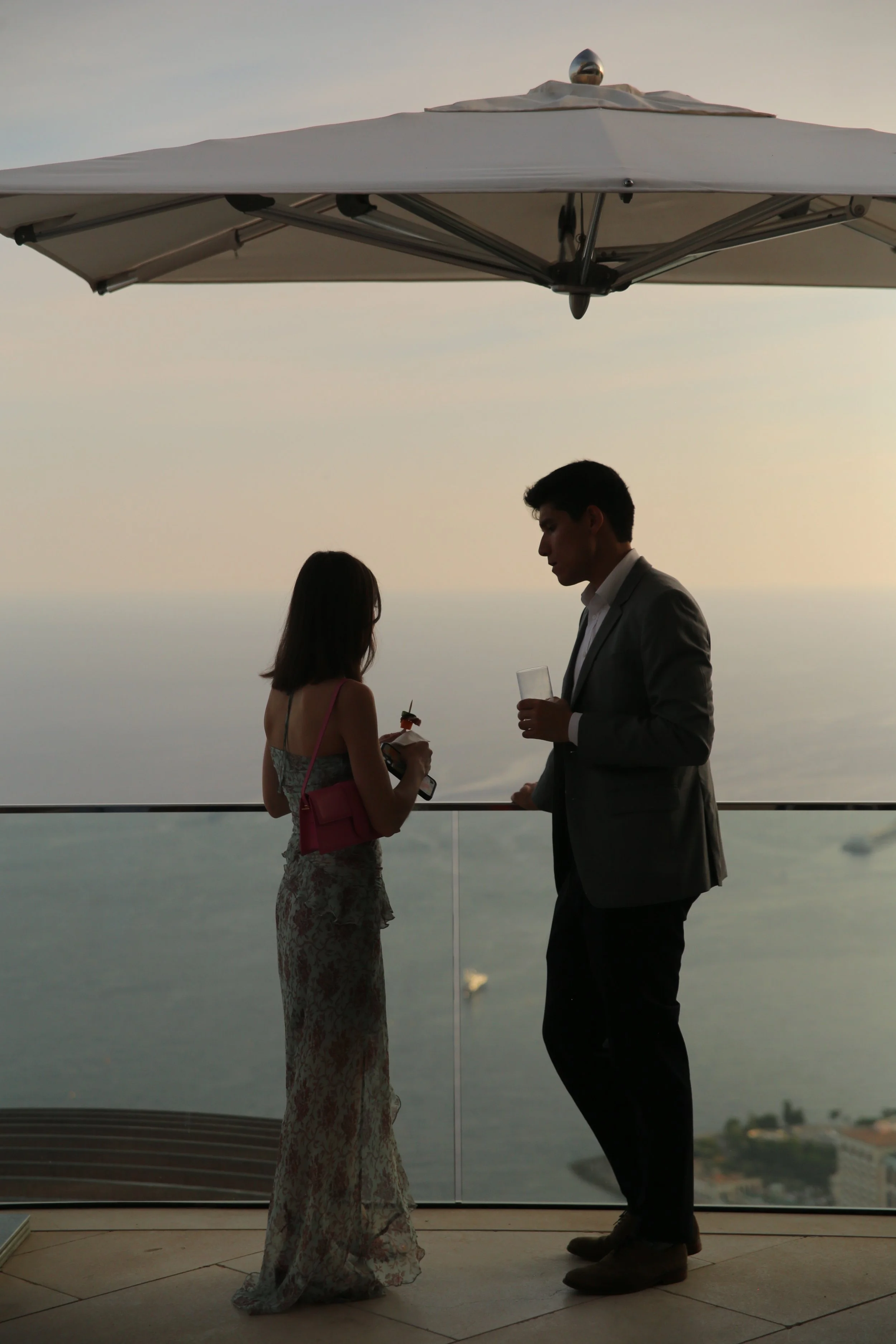 Un homme en costume et une femme en robe longues discutent sous un parasol au bord d'une terrasse avec vue sur la mer au coucher du soleil, avec un verre et une brochette dans leurs mains.