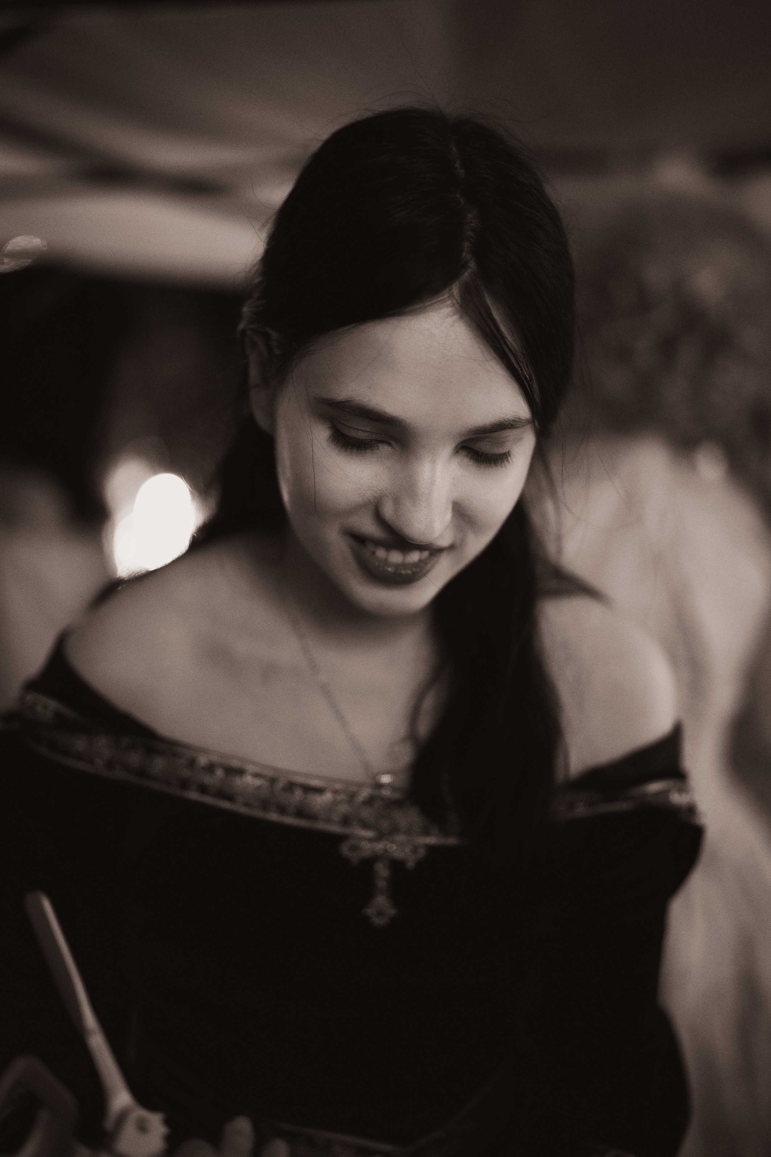 Une jeune femme avec des cheveux noirs, portant un vêtement élégant avec des motifs, sourit doucement en regardant vers le bas, dans un cadre lumineux