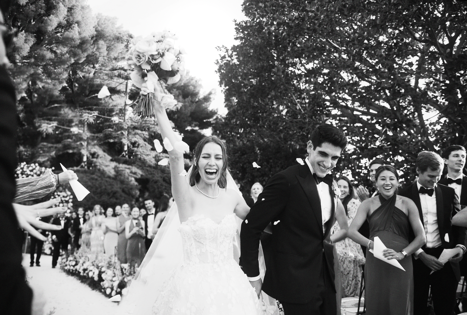 Photo en noir et blanc de jeunes mariés célébrant leur mariage, la mariée tenait un bouquet de fleurs, entourés de personnes habillées élégamment, dans un cadre extérieur avec des arbres.