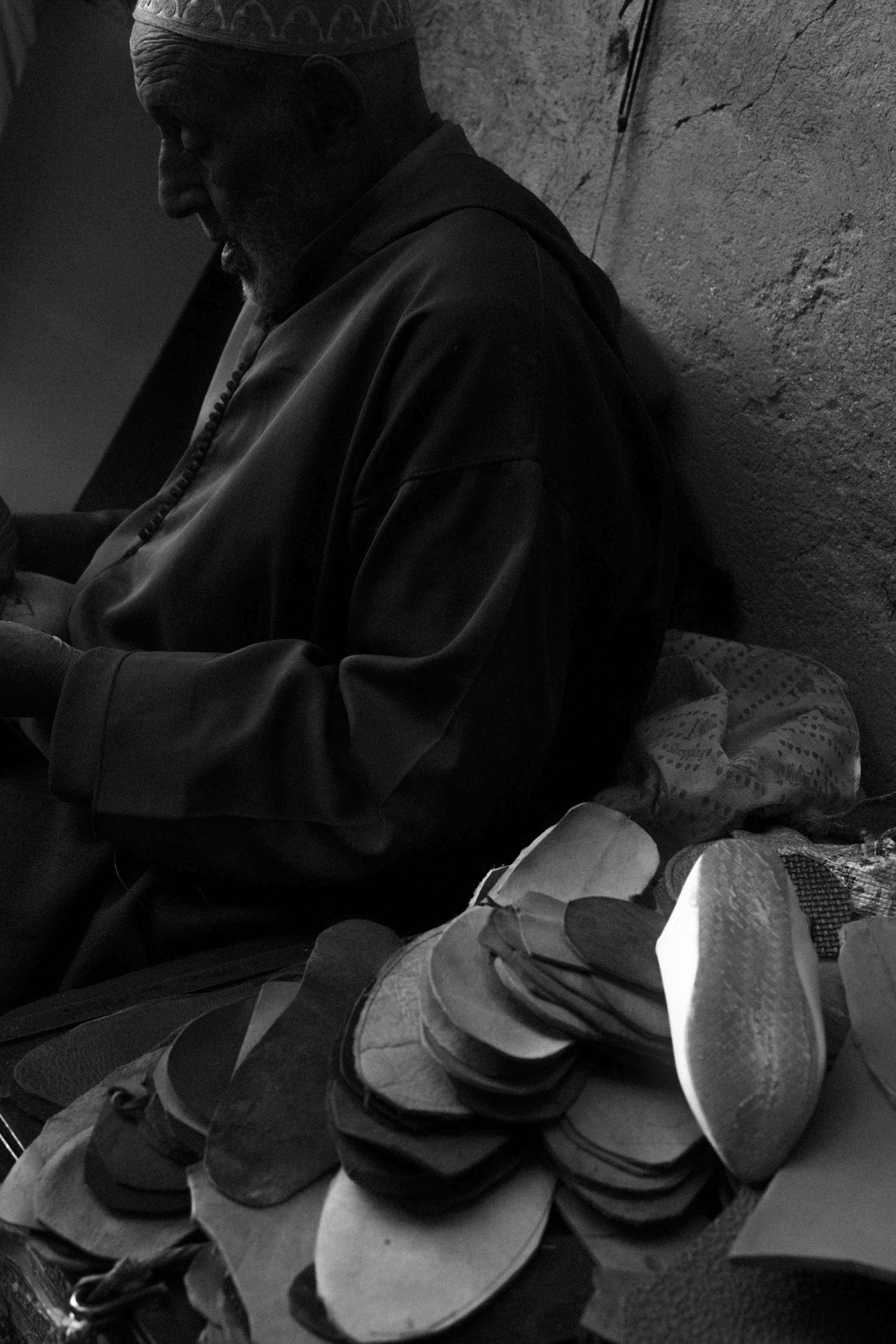 Un homme âgé portant un bonnet et une veste sombre est assis contre un mur, la tête baissée, entouré de chaussures empilées.