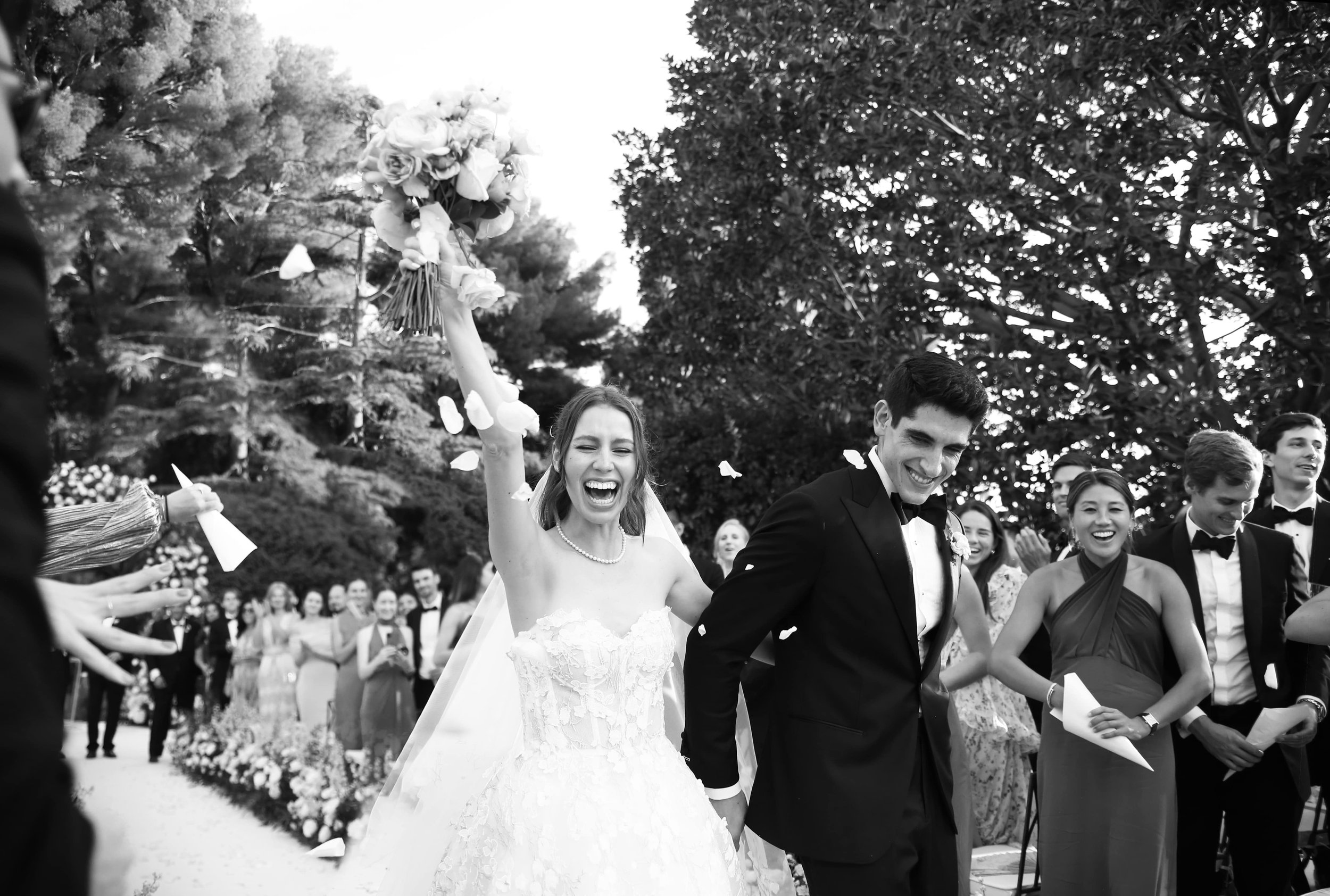 Photo d'un mariage avec des mariés souriants, la mariée en robe de mariée blanche et le marié en costume noir, entourés d'invités lors de la célébration.
