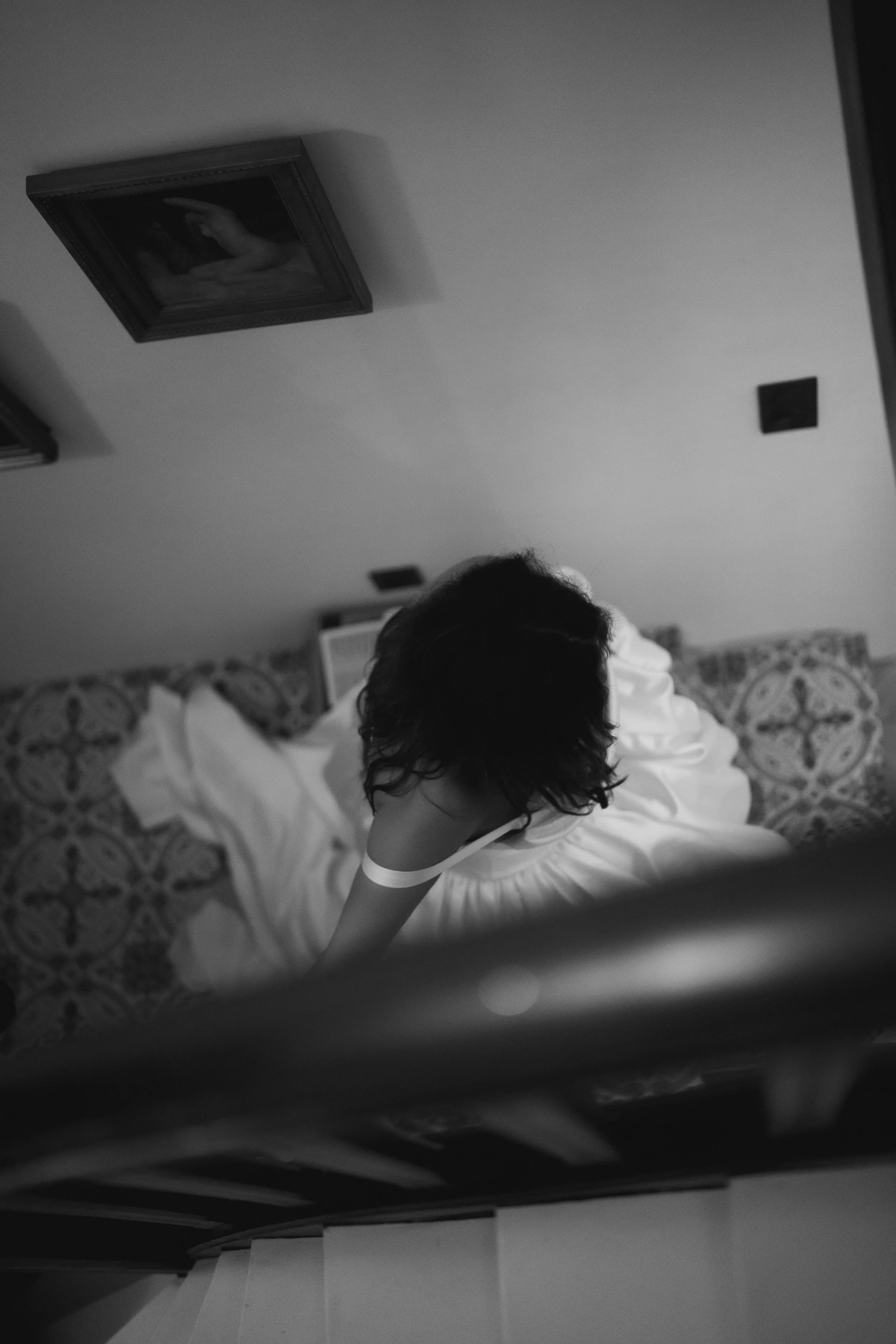 Une photographie en noir et blanc vue d'en haut d'une personne avec des cheveux longs et sombres, portant une robe blanche, assise sur un lit avec une tête reposant contre un mur décoré de motifs. La pièce comporte des œuvres d'art encadrées au plafond.