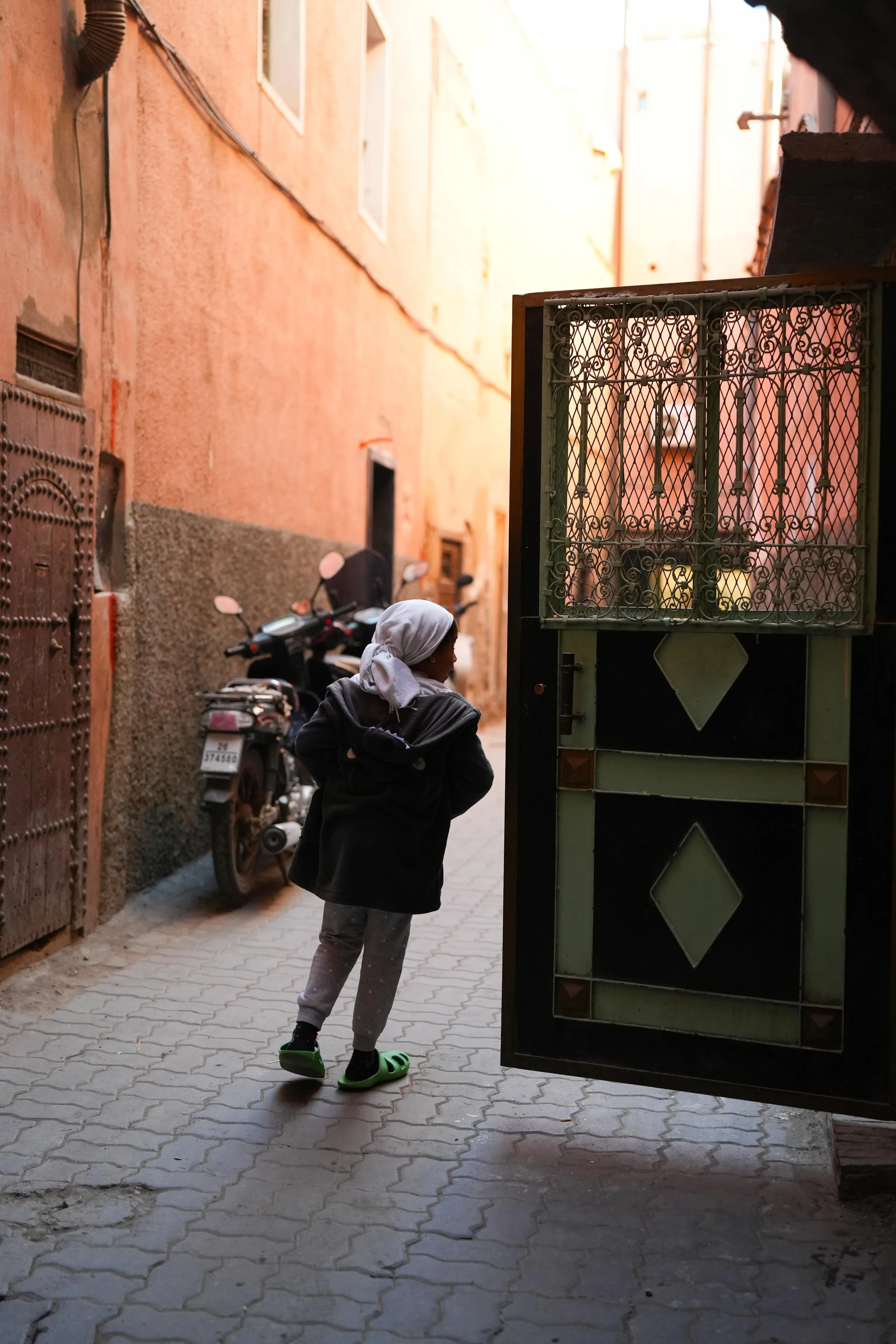 Un enfant marche dans une ruelle étroite avec des motos garées sur le côté, une porte métallique ouverte en premier-plan, murs en pierre et en plâtre aux couleurs chaudes.