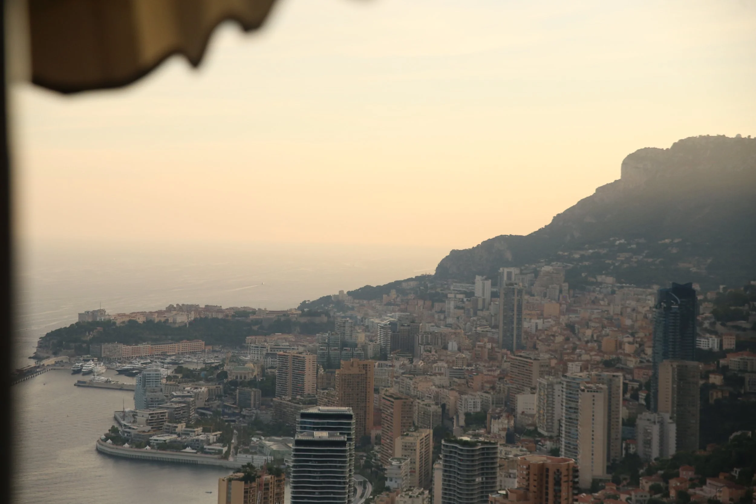 Vue aérienne de la ville de Monaco avec ses immeubles, port avec yachts, et montagne en arrière-plan, au coucher du soleil.