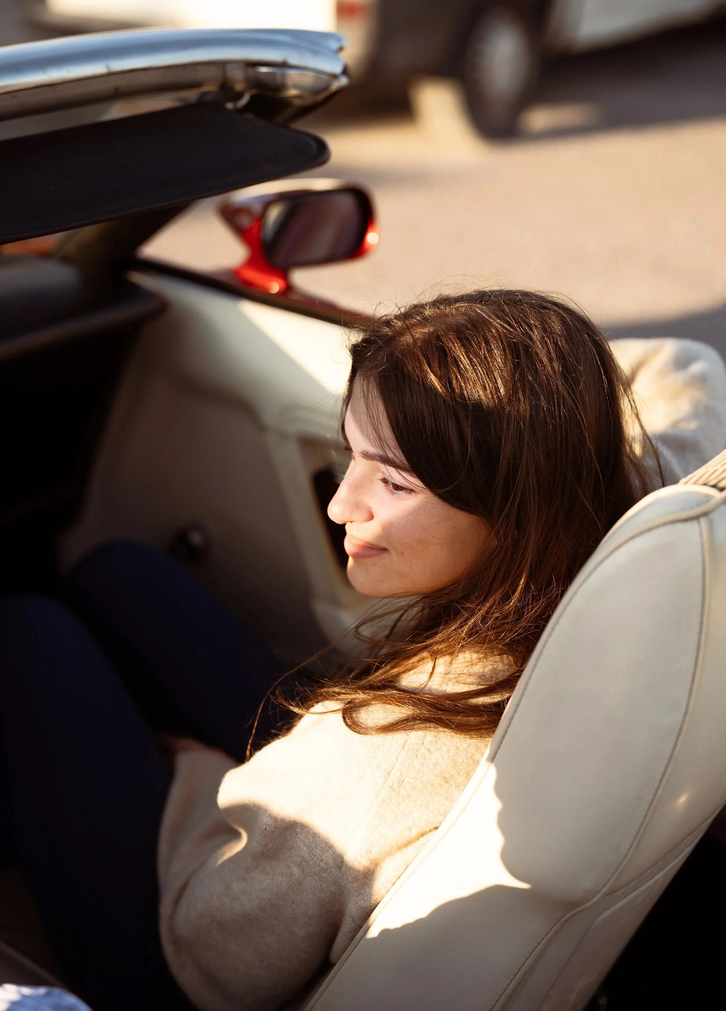 Femme assise dans une voiture, profitant du soleil, avec des cheveux bruns et un manteau beige.