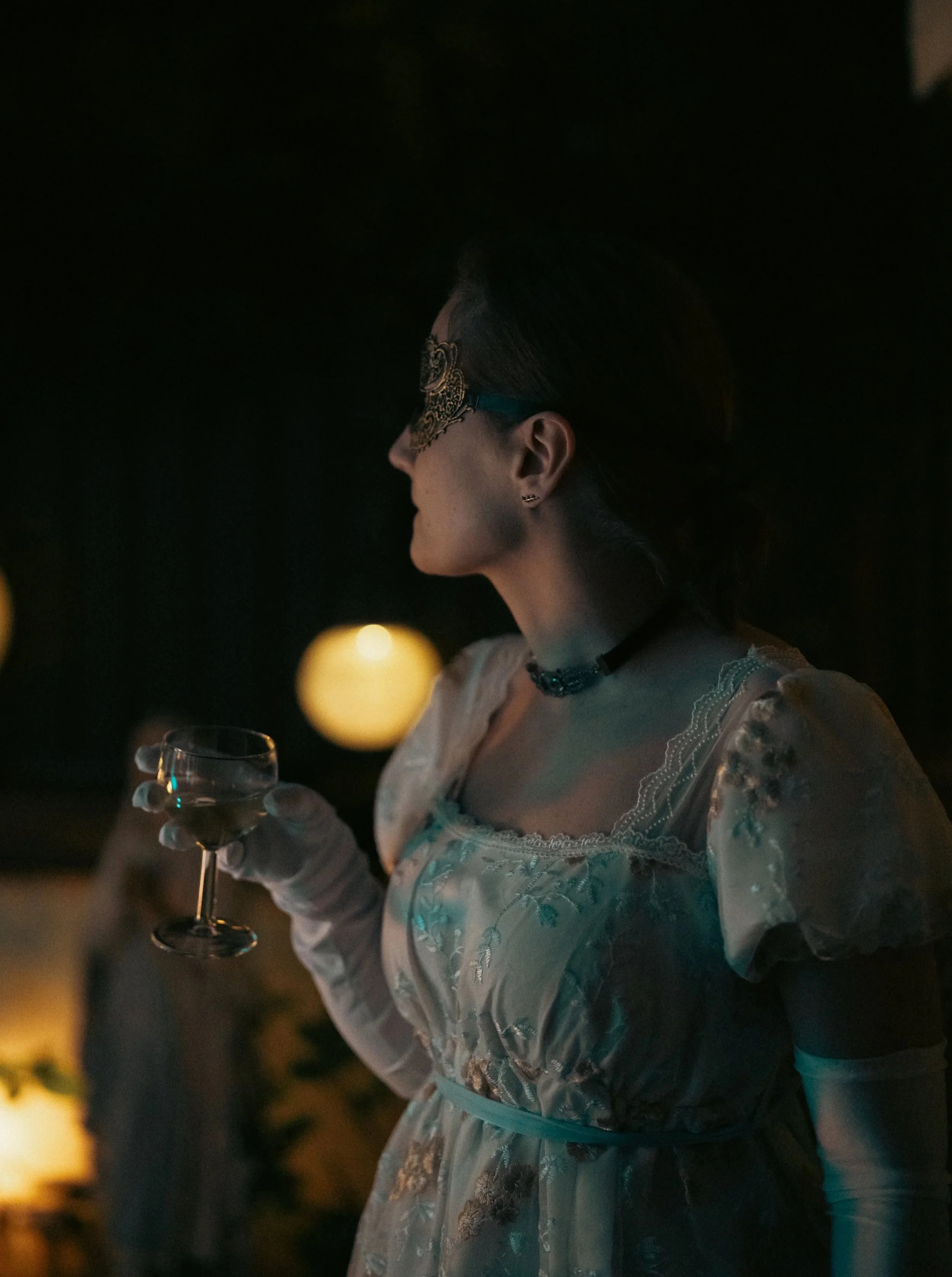 Femme portant une robe élégante blanche avec dentelle et gants blancs, tenant un verre à cocktail, dans un cadre sombre avec des lumières chaudes en arrière-plan, portant un masque de carnaval doré.