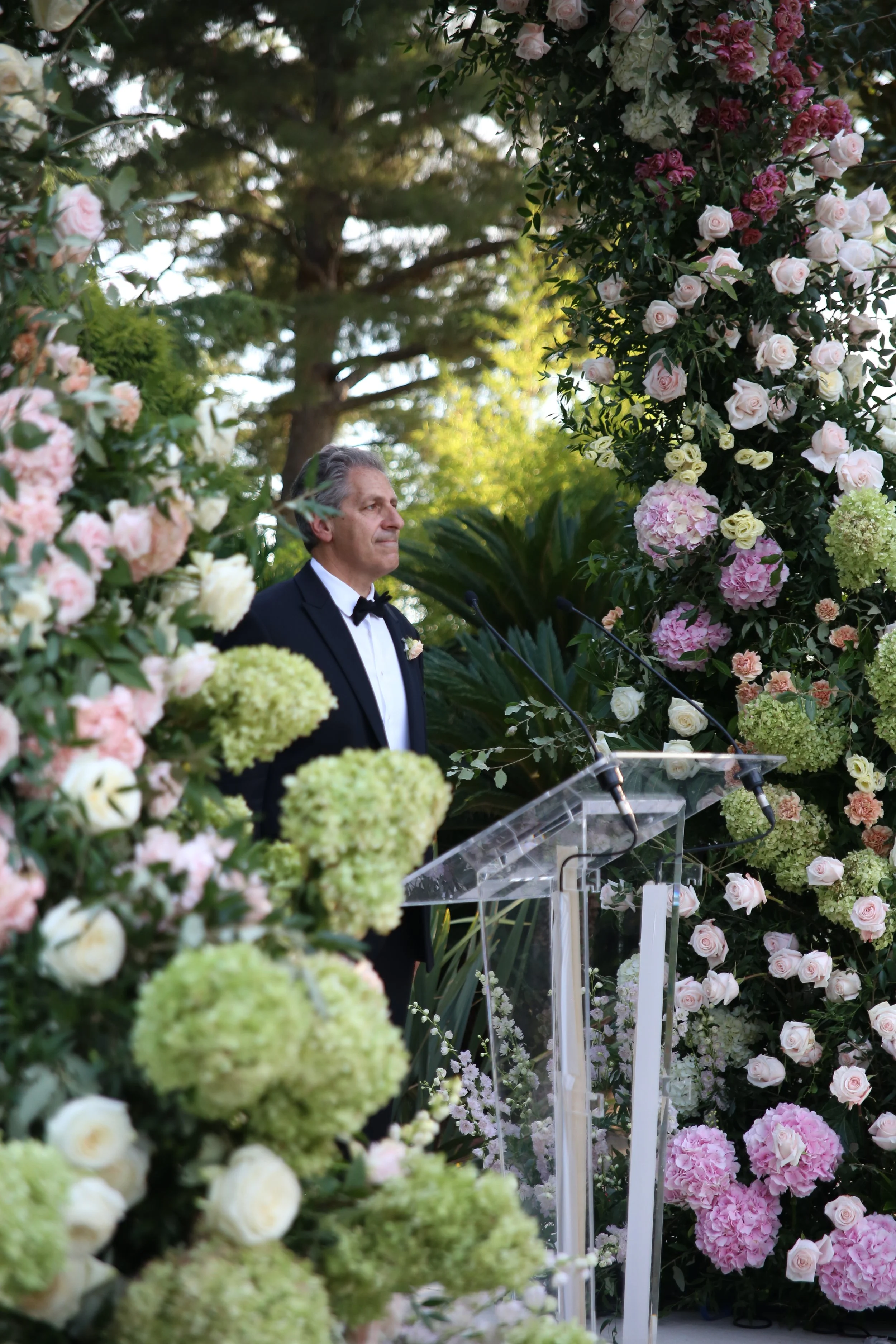 Un homme en smoking donnant un discours lors d'une cérémonie, entouré de fleurs de roses et d'hortensias roses et vertes, en extérieur avec des arbres en fond.