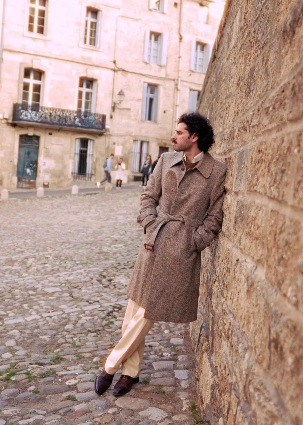 Un homme avec des cheveux bouclés et une moustache, portant un long manteau marron, se tient contre un mur en pierre sur une place pavée de ville, avec des bâtiments en arrière-plan.