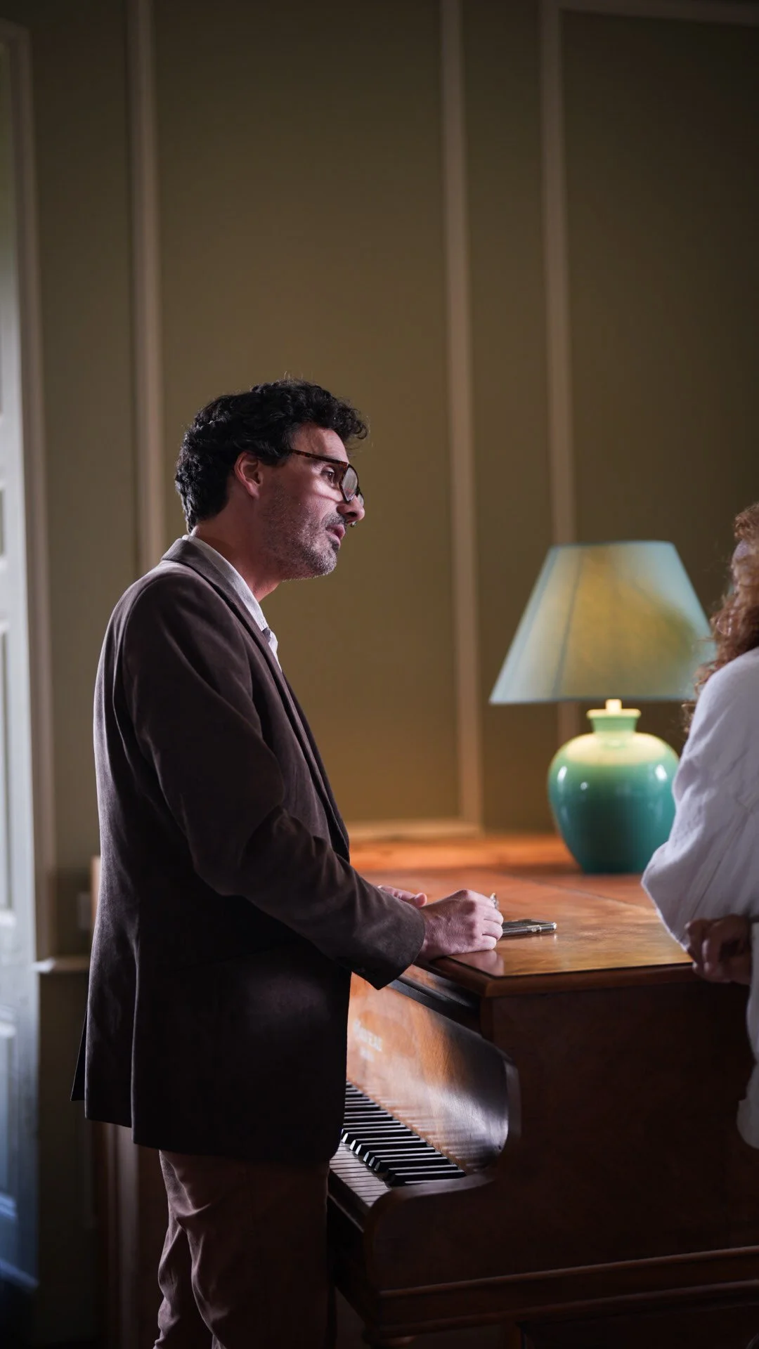 Homme avec lunettes et barbe, en costume marron, debout près d'un piano en bois, dans une pièce avec une lampe verte en céramique sur une table à côté.