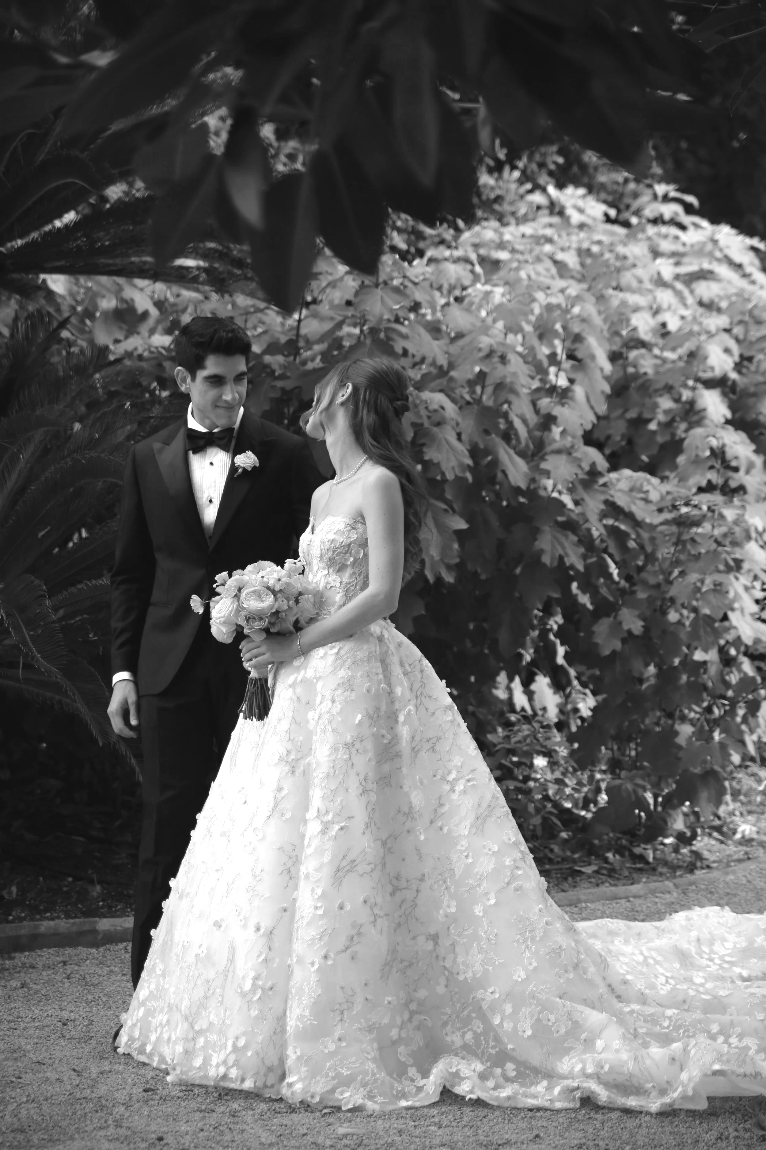 Un couple en mariage, habillé élégamment, se regarde tendrement dans un jardin avec de grands feuillages en arrière-plan.
