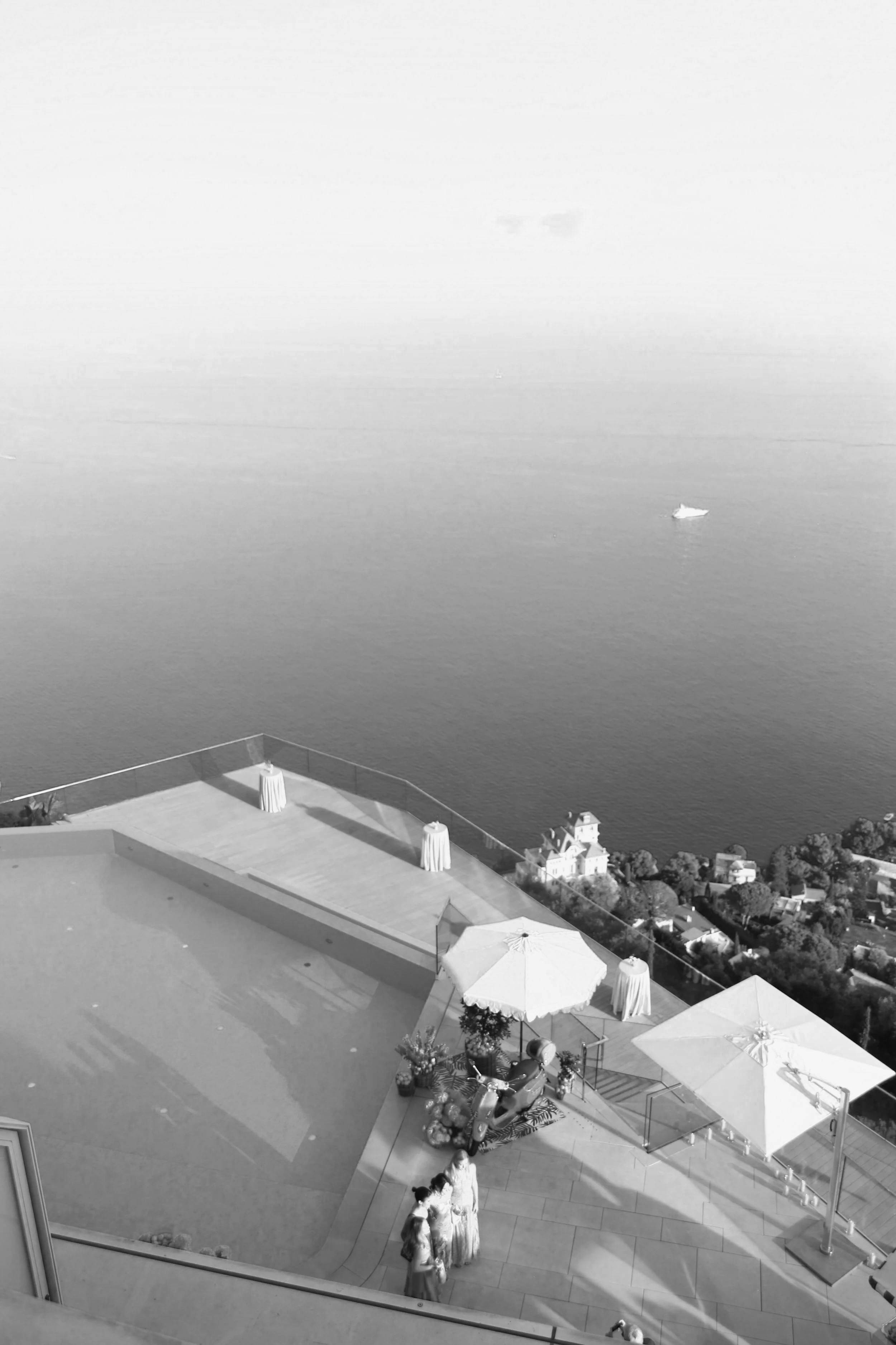 Vue d'une terrasse en hauteur avec parasols, une moto, et des personnes, surplombant la mer avec un bateau, en noir et blanc.