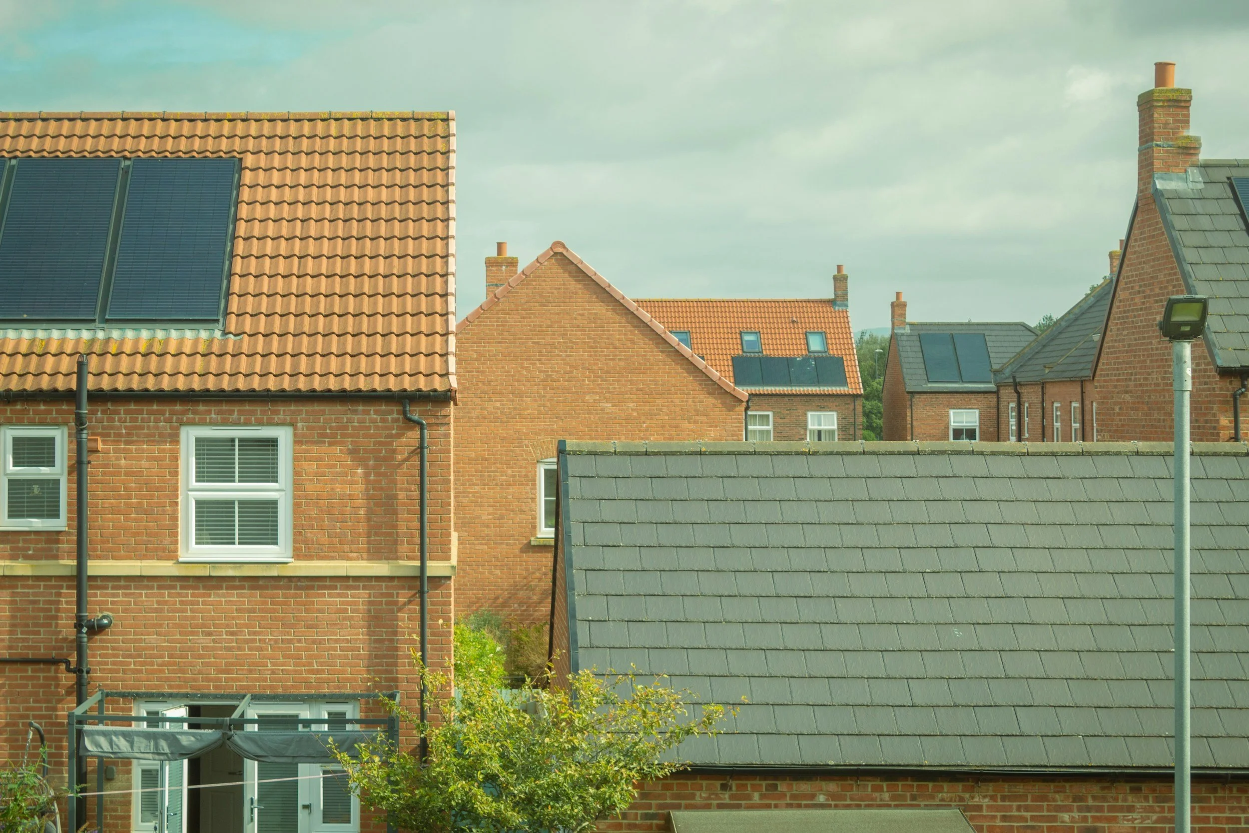 Multiple residential houses with brick walls, tiled and shingle roofs, and solar panels, under a cloudy sky.