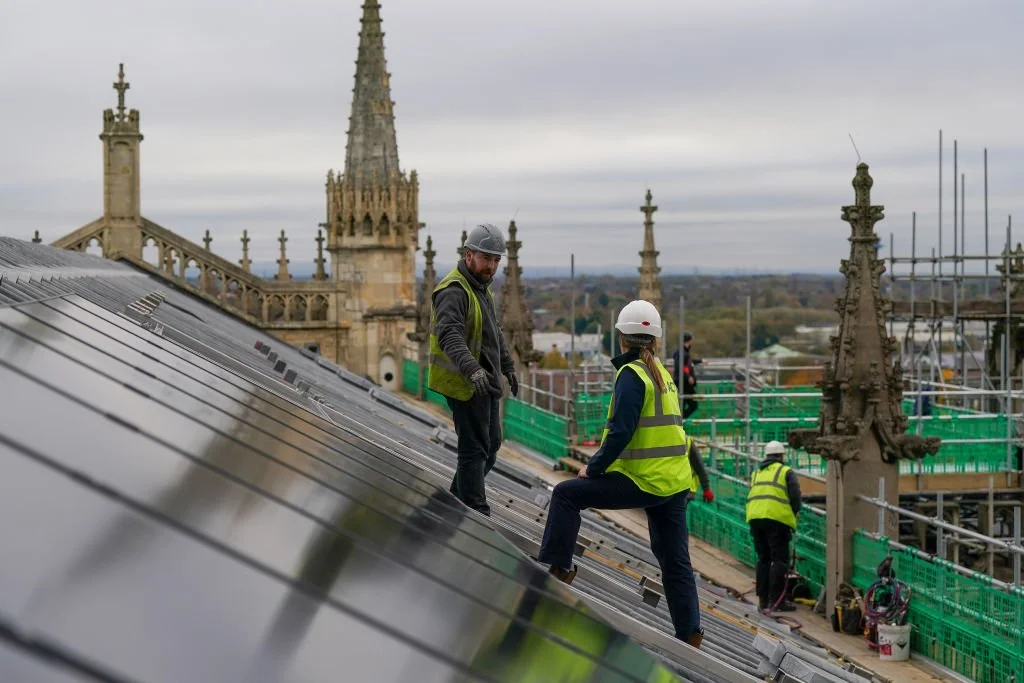 Construction workers wearing safety vests and hard hats on the roof of a building with gothic architecture, scaffolding, and cityscape in the background.