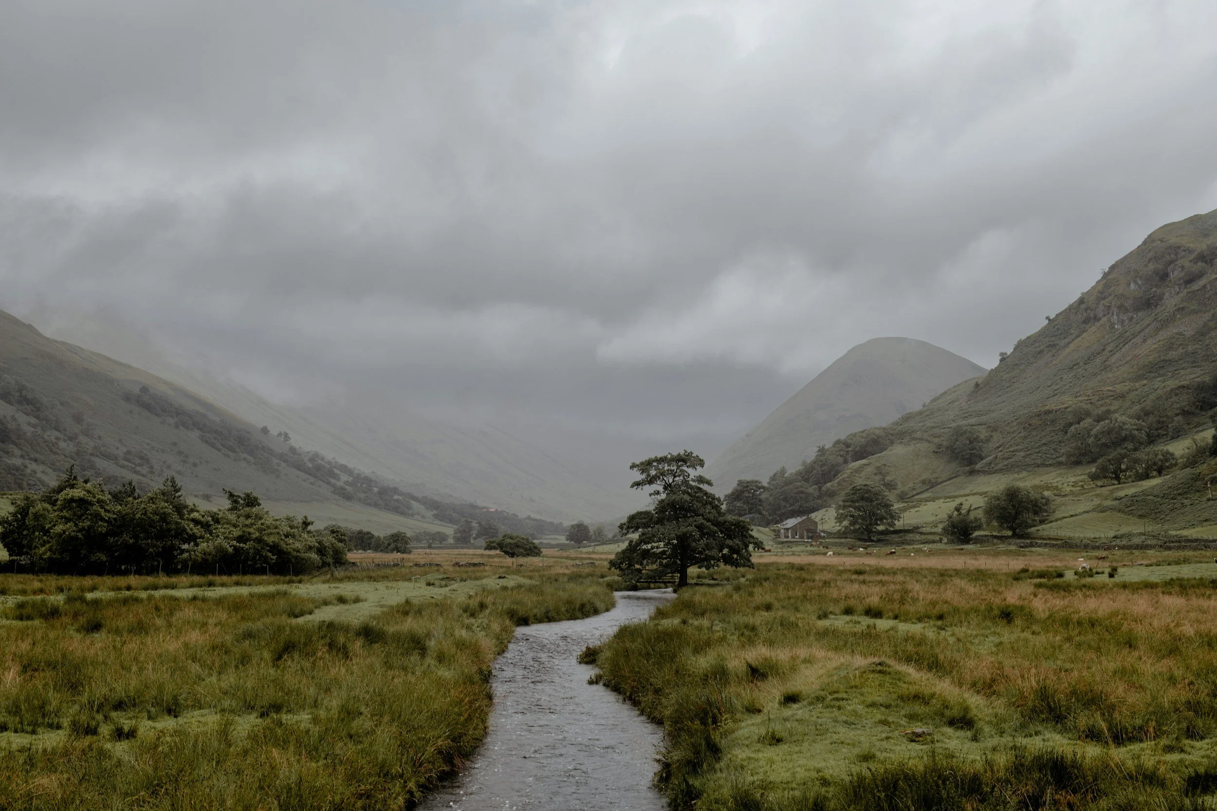 A scenic valley with a small river flowing through grassy land, surrounded by rolling hills and mountains under a cloudy sky.