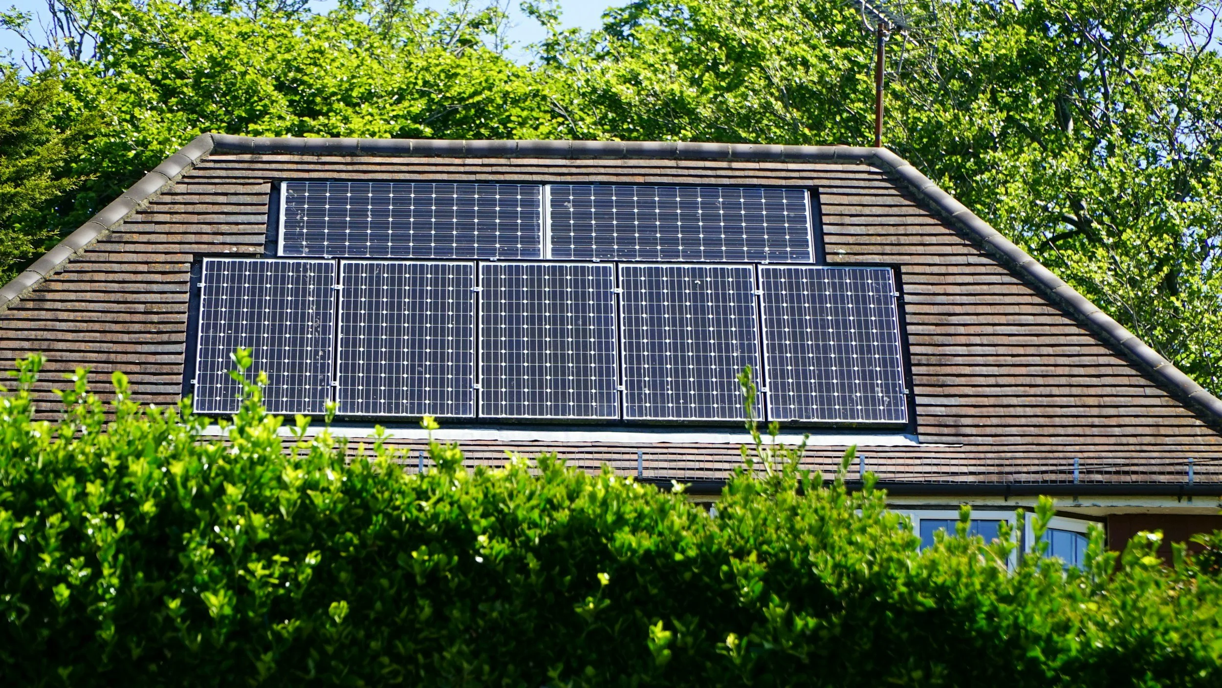House roof with solar panels, surrounded by green bushes and trees in the background.