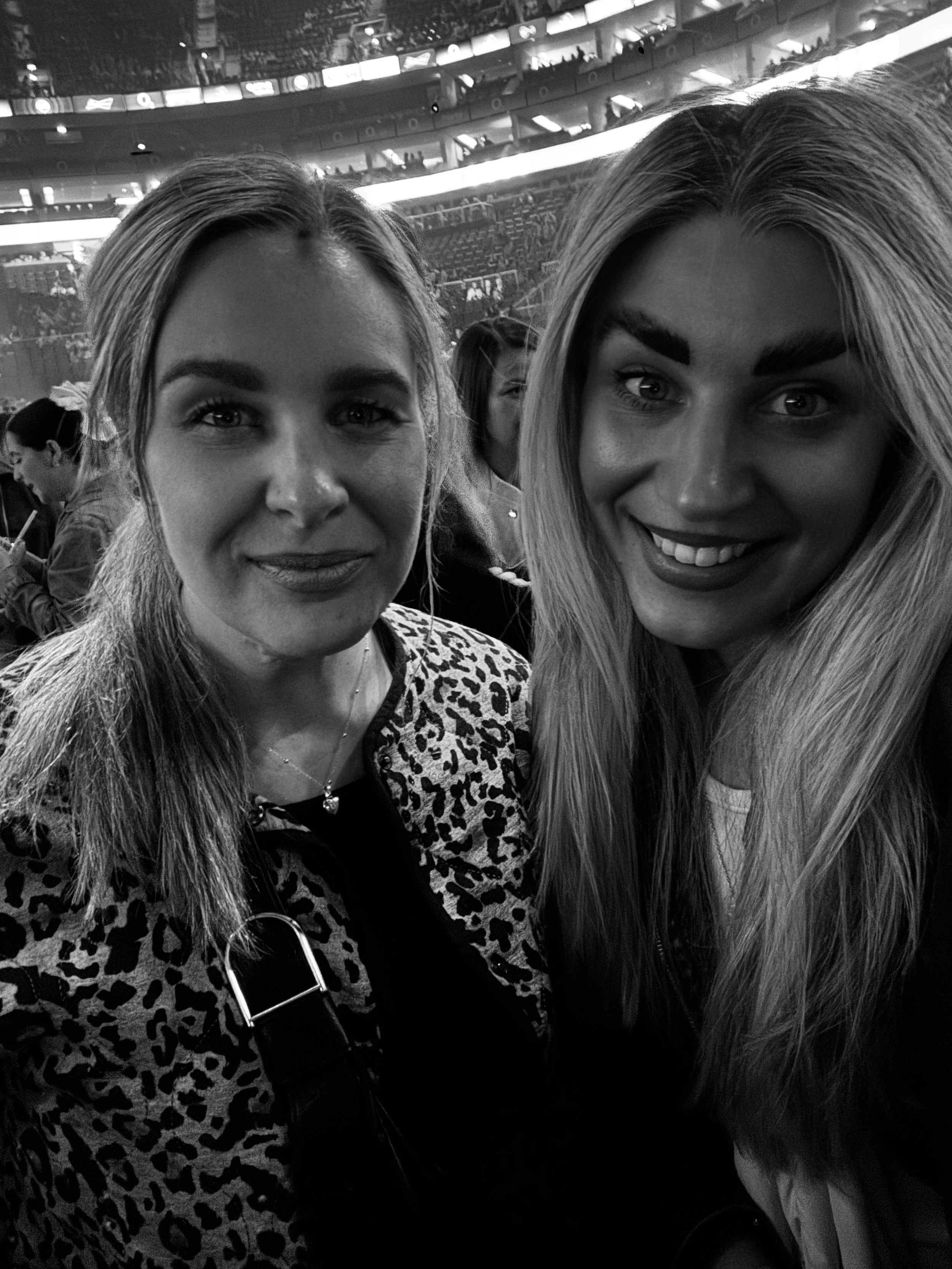 Two women taking a selfie at an indoor event or concert with a large audience in the background.