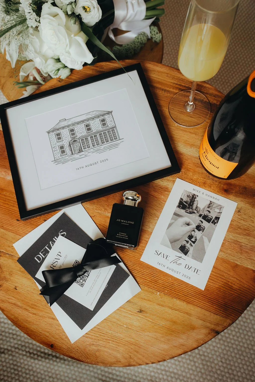 A wooden table with a framed artistic drawing of a house and the date 16th August 2025, a bottle of Jo Malone perfume, a black and yellow champagne bottle, a glass of champagne, a wedding invitation with a black bow, a black and white photograph print, and a floral bouquet in the background.