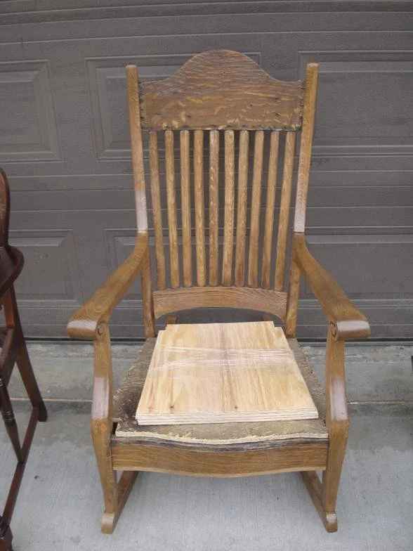 A wooden chair with a high backrest, armrests, and slats, missing its seat cushion, with a piece of plywood placed on the seat area, positioned in front of a garage door.