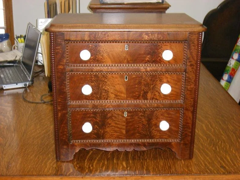 A vintage wooden three-drawer dresser with round keyholes and decorative trim, placed on a wooden table.