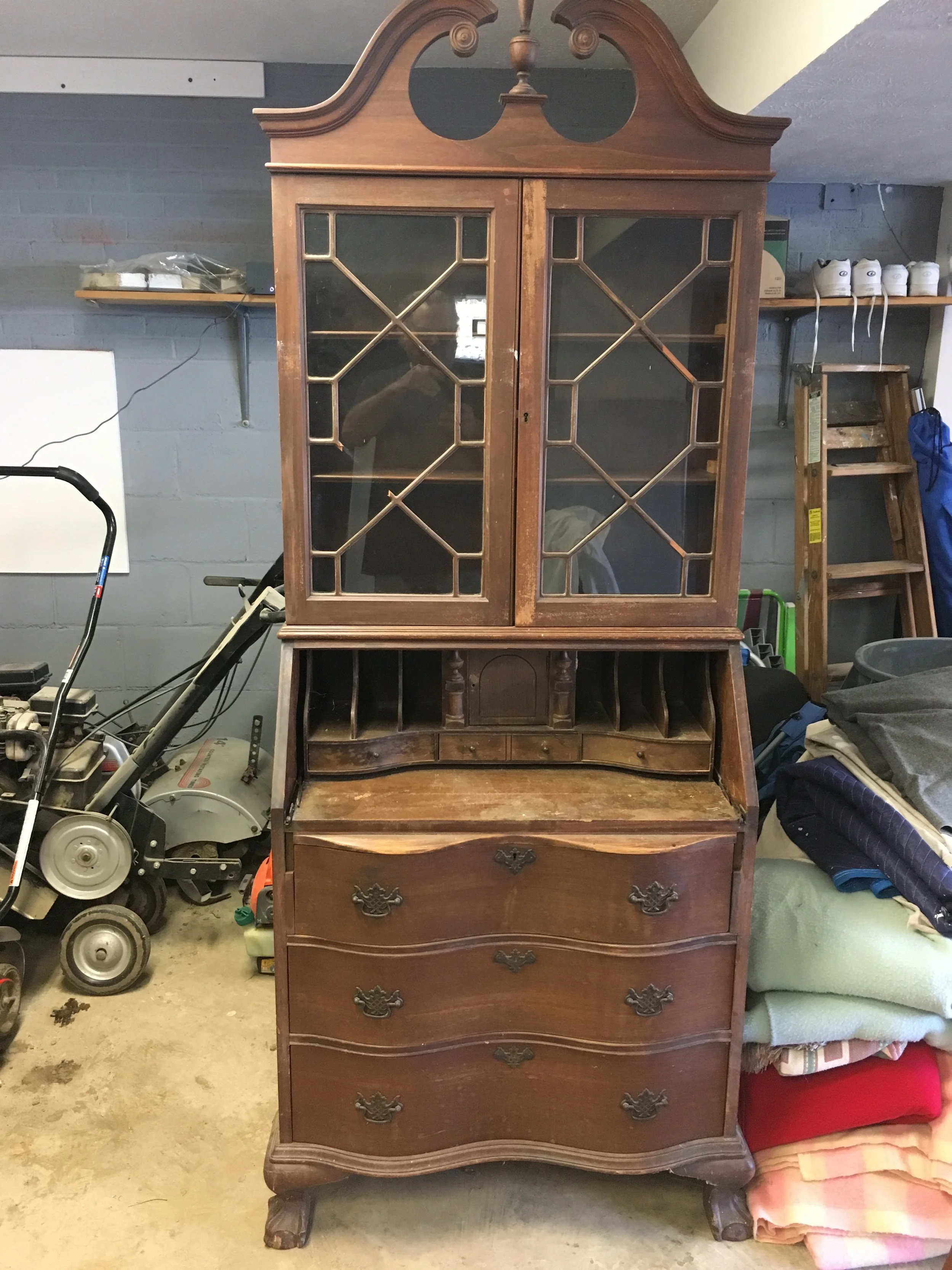 An antique wooden hutch with glass-paneled upper doors, a small writing desk section with curved drawers in the middle, and three larger drawers below, situated in a cluttered garage with various tools, stacked blankets, and shelving in the background.