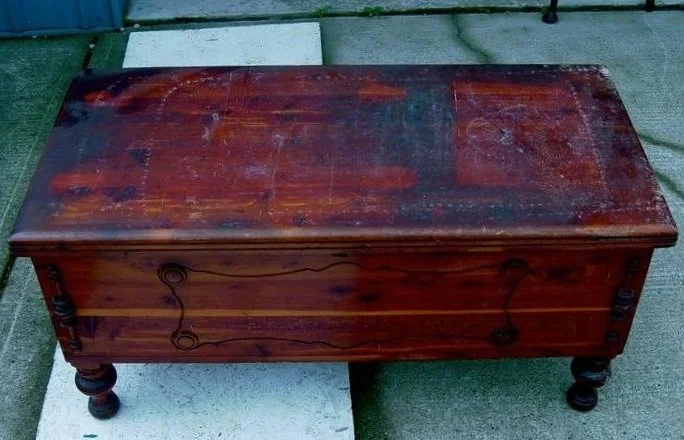 A wooden chest or storage box with a worn, reddish-brown finish and turned legs, placed outdoors on a sidewalk.