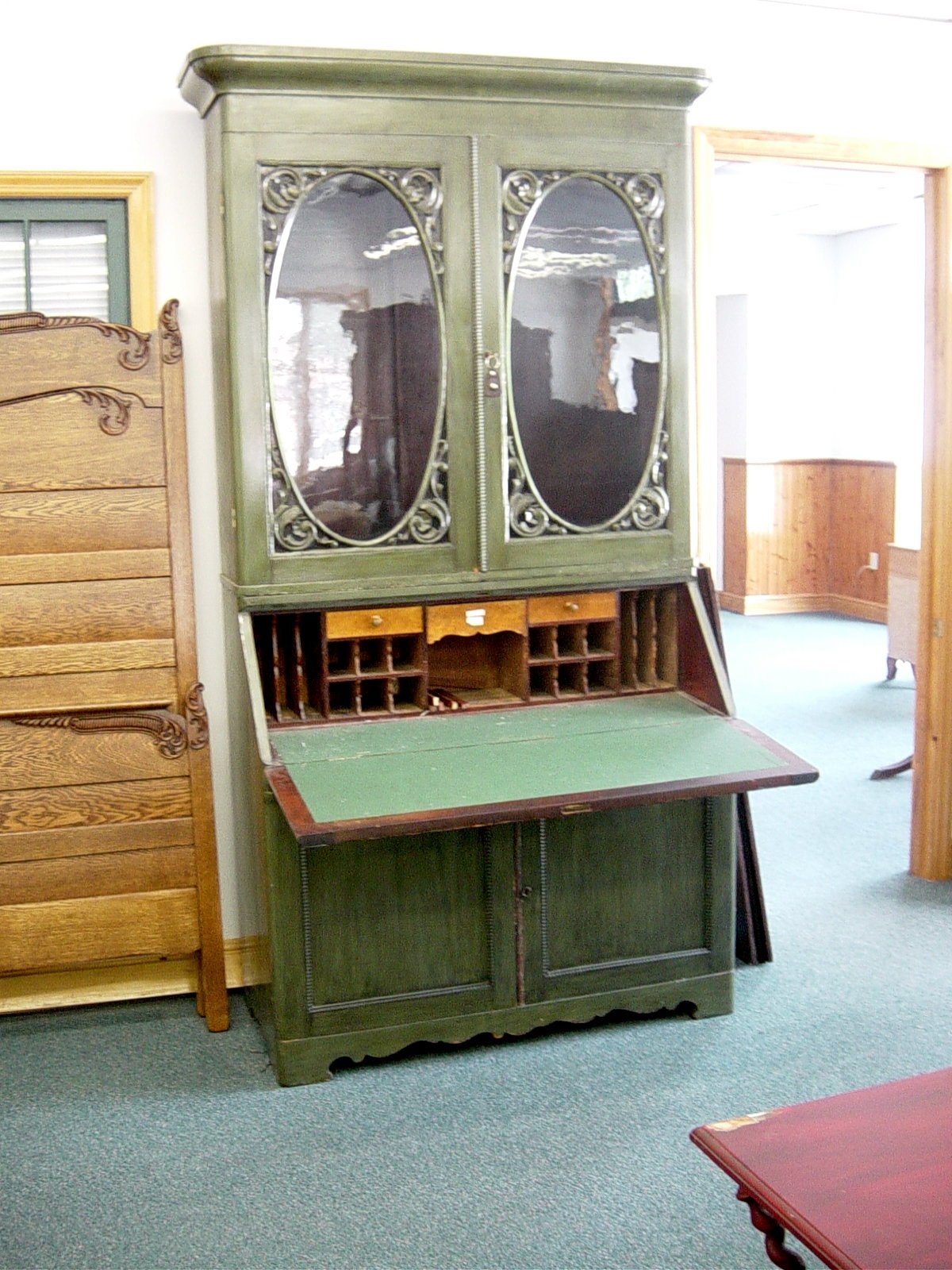 A vintage green secretary desk with built-in glass-front cabinets and small compartments, located in a room with wooden furniture and green carpet.