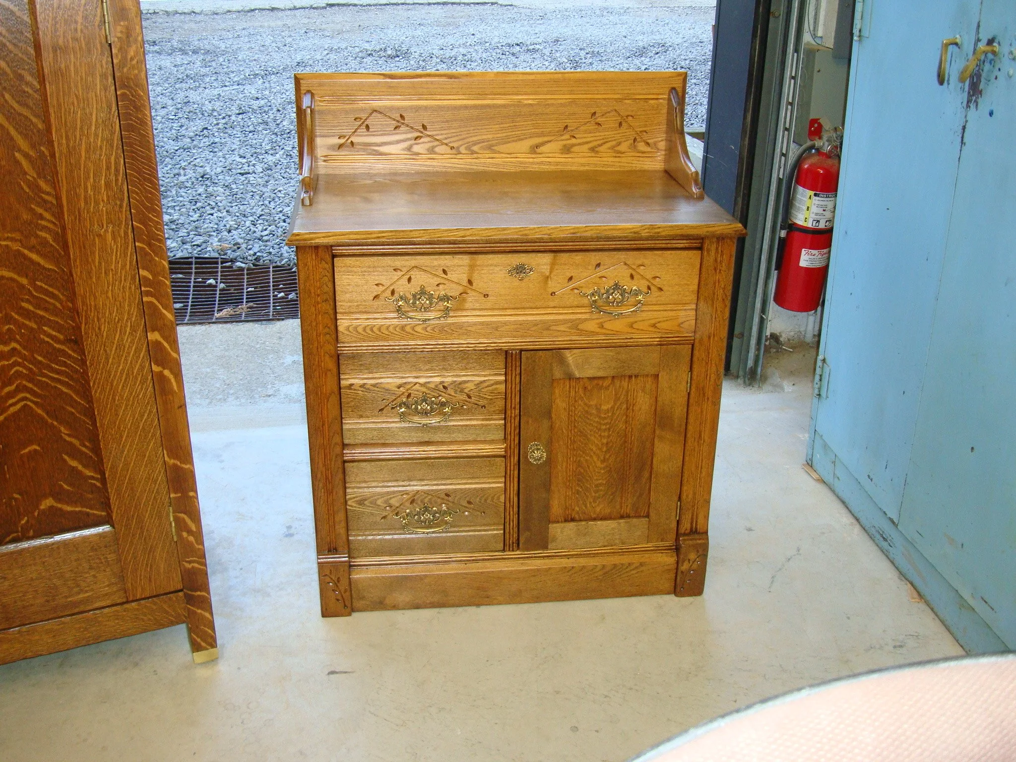 A small wooden cabinet with decorative brass hardware and a folding top, placed on a concrete floor near a blue wall and a fire extinguisher.
