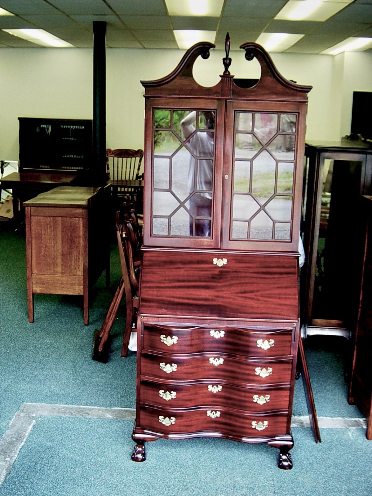 A vintage wooden secretary bureau with a glass-paneled upper cabinet and ornate brass handles, standing on small carved feet, in a room with other wood furniture pieces.