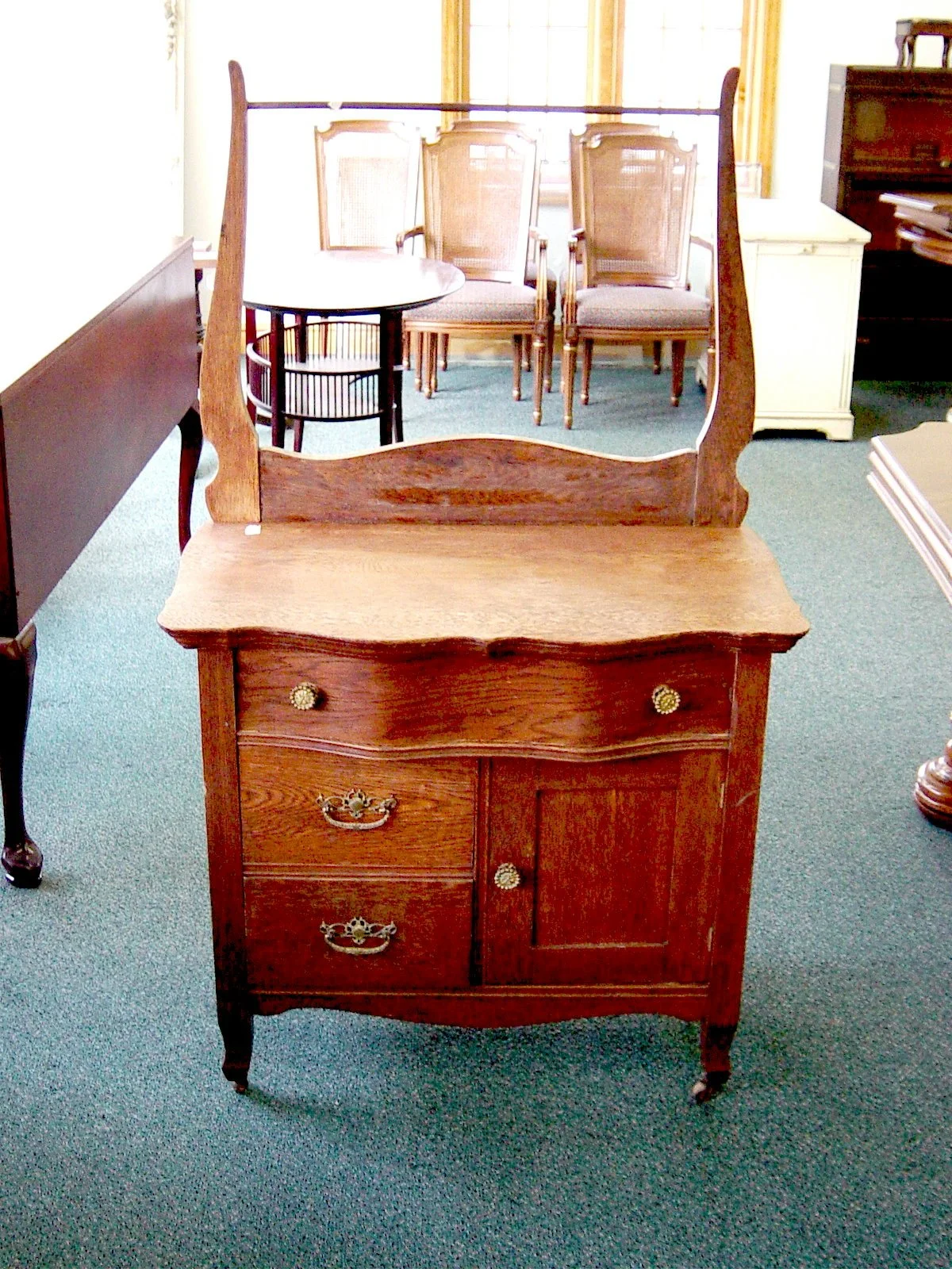Antique wooden dresser with mirror, three small drawers, one cabinet, and decorative metal handles, standing on a green carpet in a room with chairs and other furniture.
