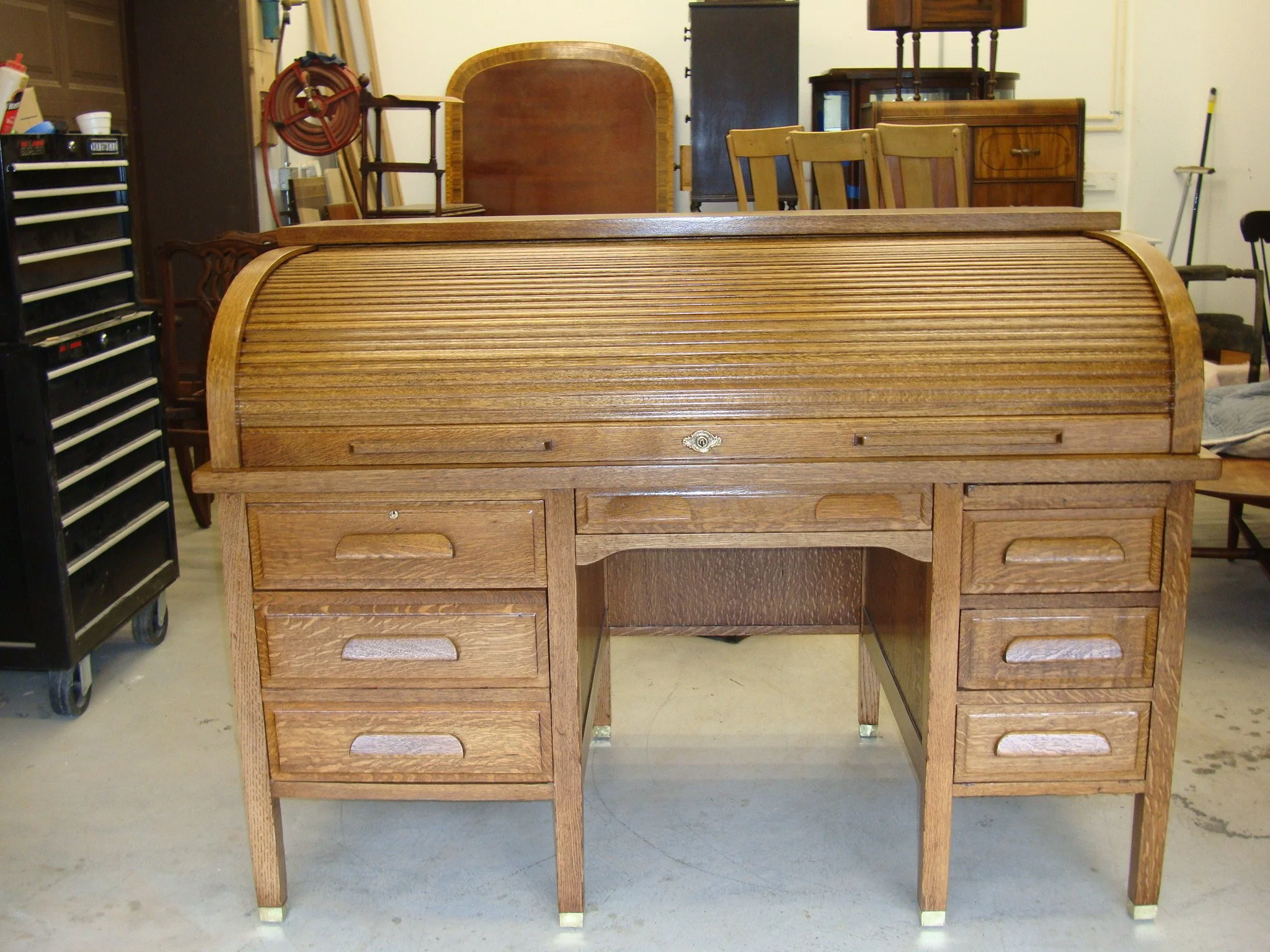 A vintage roll-top wooden desk with multiple drawers on either side, situated in a workshop or storage area with various furniture and tools in the background.