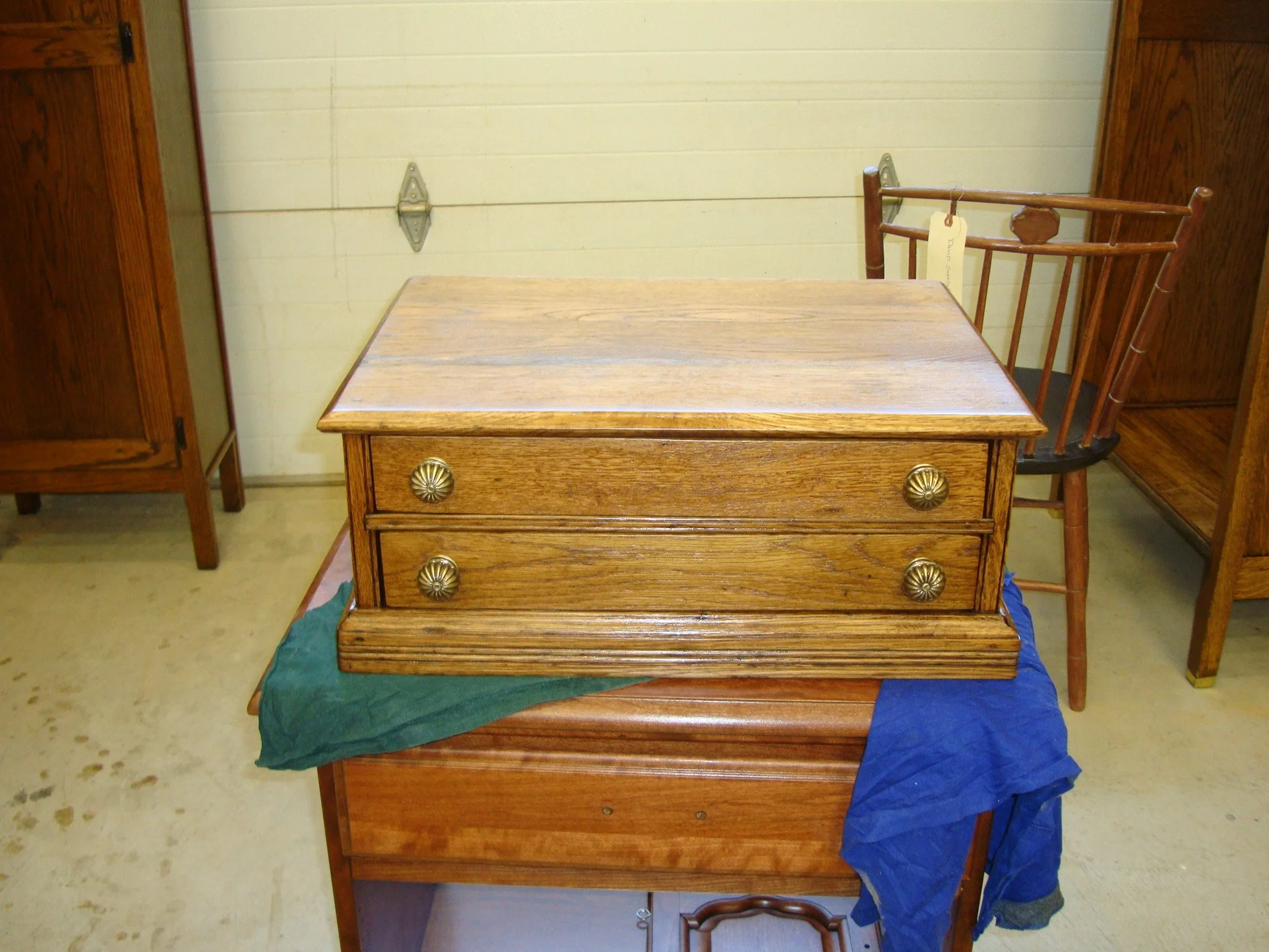 A wooden jewelry box with two drawers, brass-colored knobs, and a flat top, placed on a green cloth and blue cloth on a small table. In the background, there are wooden cabinets and a white garage door.