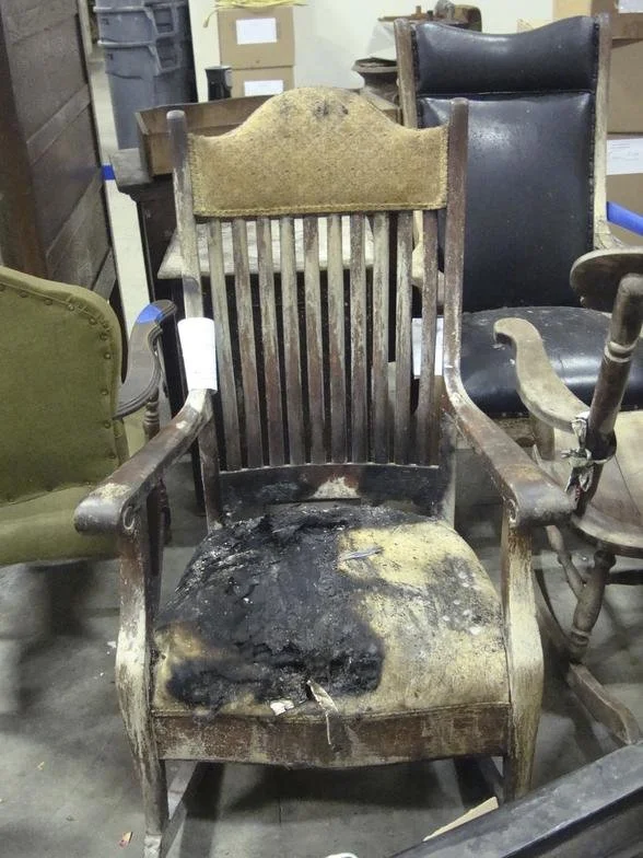 A wooden armchair with a damaged, burnt seat and backrest, surrounded by other vintage chairs in a storage or antique shop.