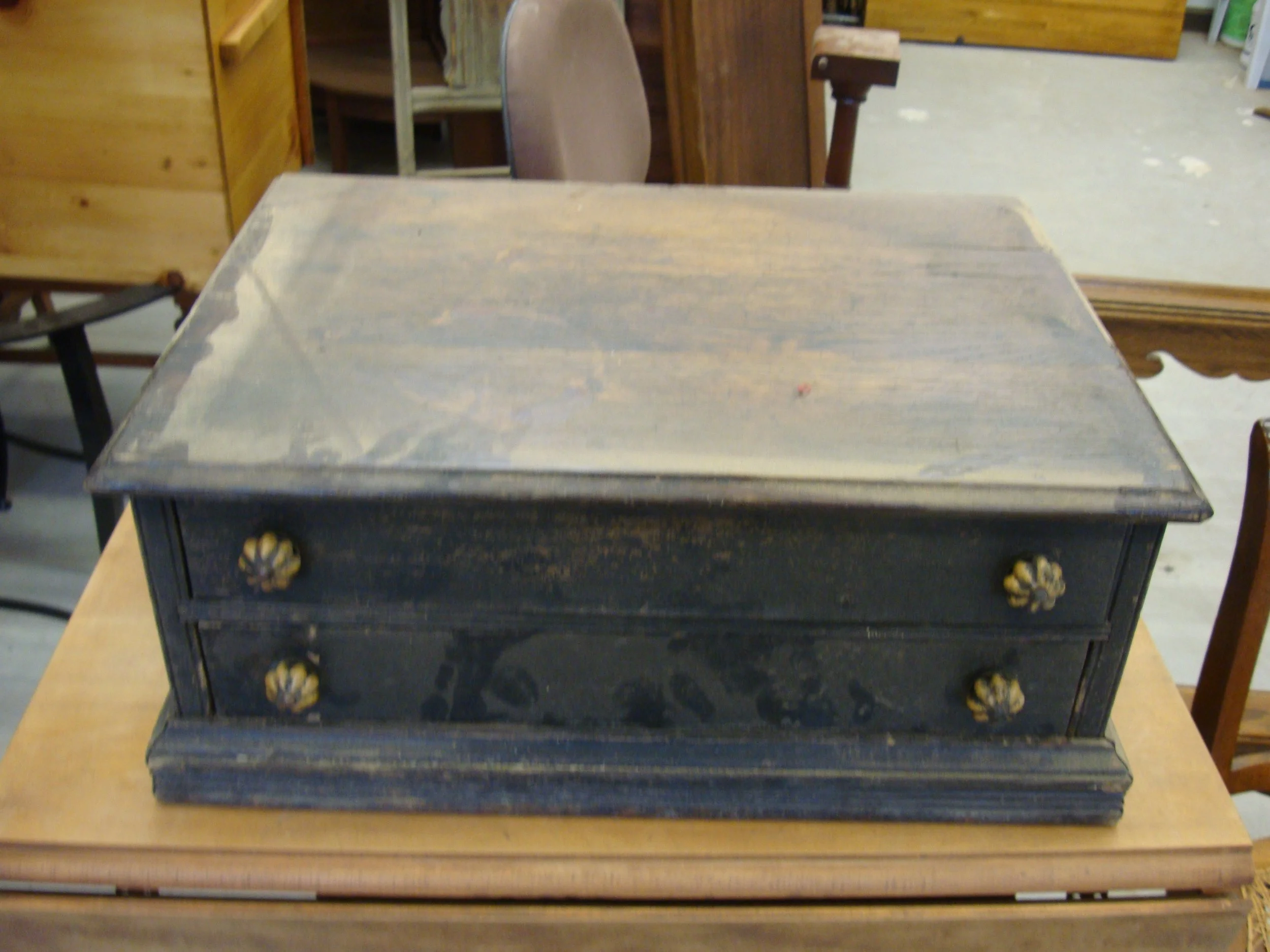 Vintage wooden jewelry box with two drawers and decorative knobs, showing signs of age and wear, placed on a wooden table in a furniture store.