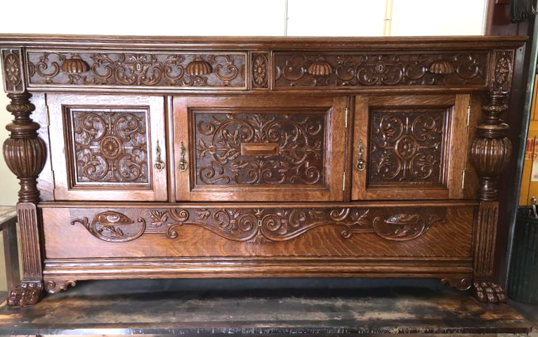 Ornate vintage wooden sideboard with carved floral and scroll design, featuring columns on the sides, three cabinet doors, and a carved apron.