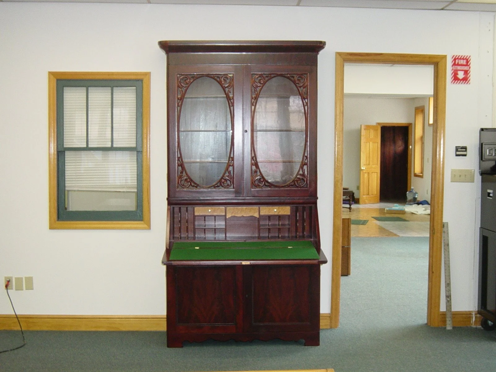 A wooden display cabinet with glass doors, a green felt writing surface, and small compartments at the bottom, placed against a white wall in a room with carpeted floor.