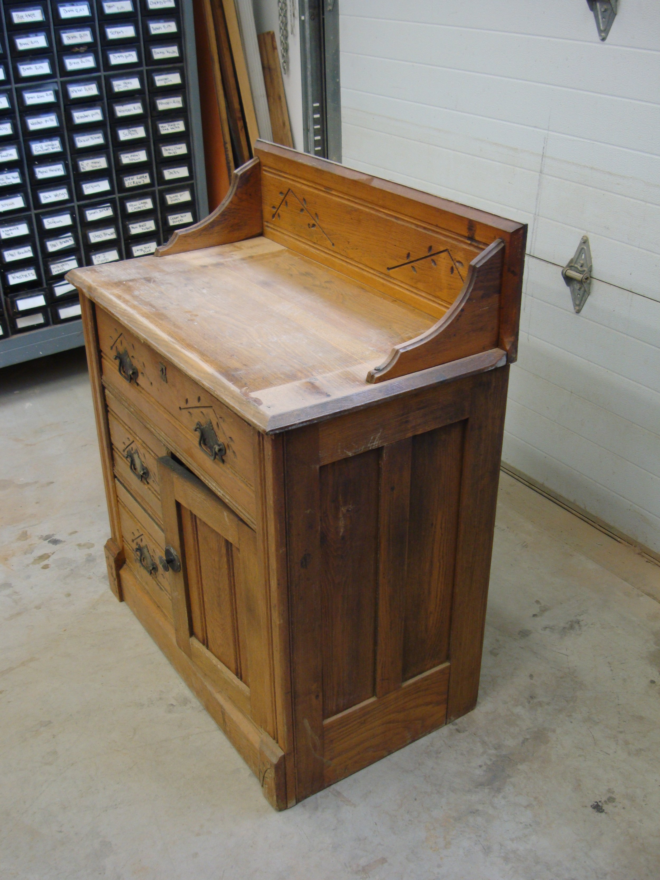 A vintage wooden kitchen cabinet with a raised back and decorative metal hardware in a workshop or garage setting.